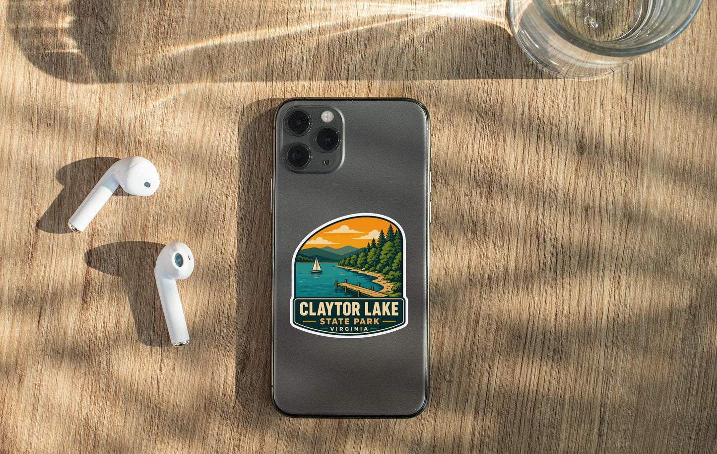a smartphone with a sticker of Claytor Lake State Park on its back, along with a pair of white earbuds and a glass of water on a wooden surface.