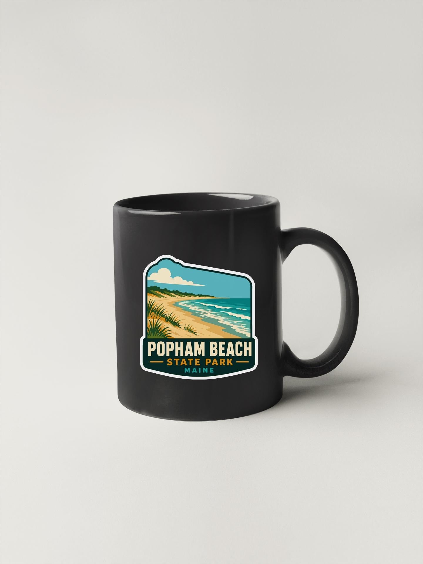 A black coffee mug with a sticker depicting a beach scene and the text "POPHAM BEACH STATE PARK" printed on it.