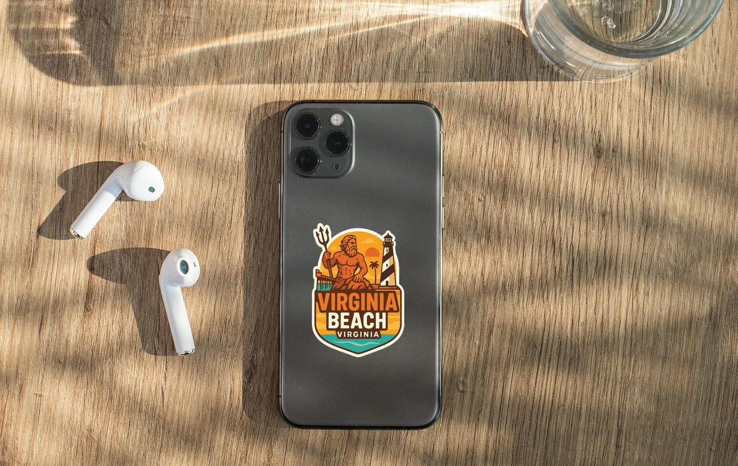 a smartphone with a sticker on the back, placed on a wooden surface alongside a pair of white earbuds and a glass of water.