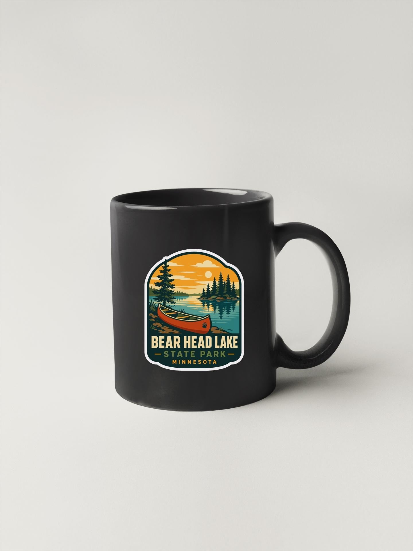 A black coffee mug with a colorful graphic design depicting a canoe on Bear Head Lake, surrounded by trees and a sunset.