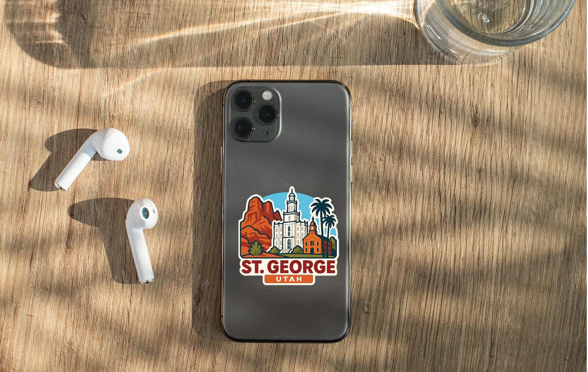 a smartphone with a sticker of St. George, Utah, placed on a wooden surface alongside a pair of white earbuds and a glass of water.