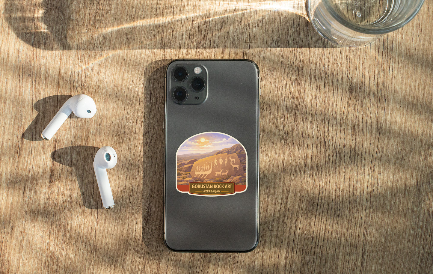 a smartphone with a sticker on the back, placed on a wooden surface alongside a pair of white earbuds and a glass of water.
