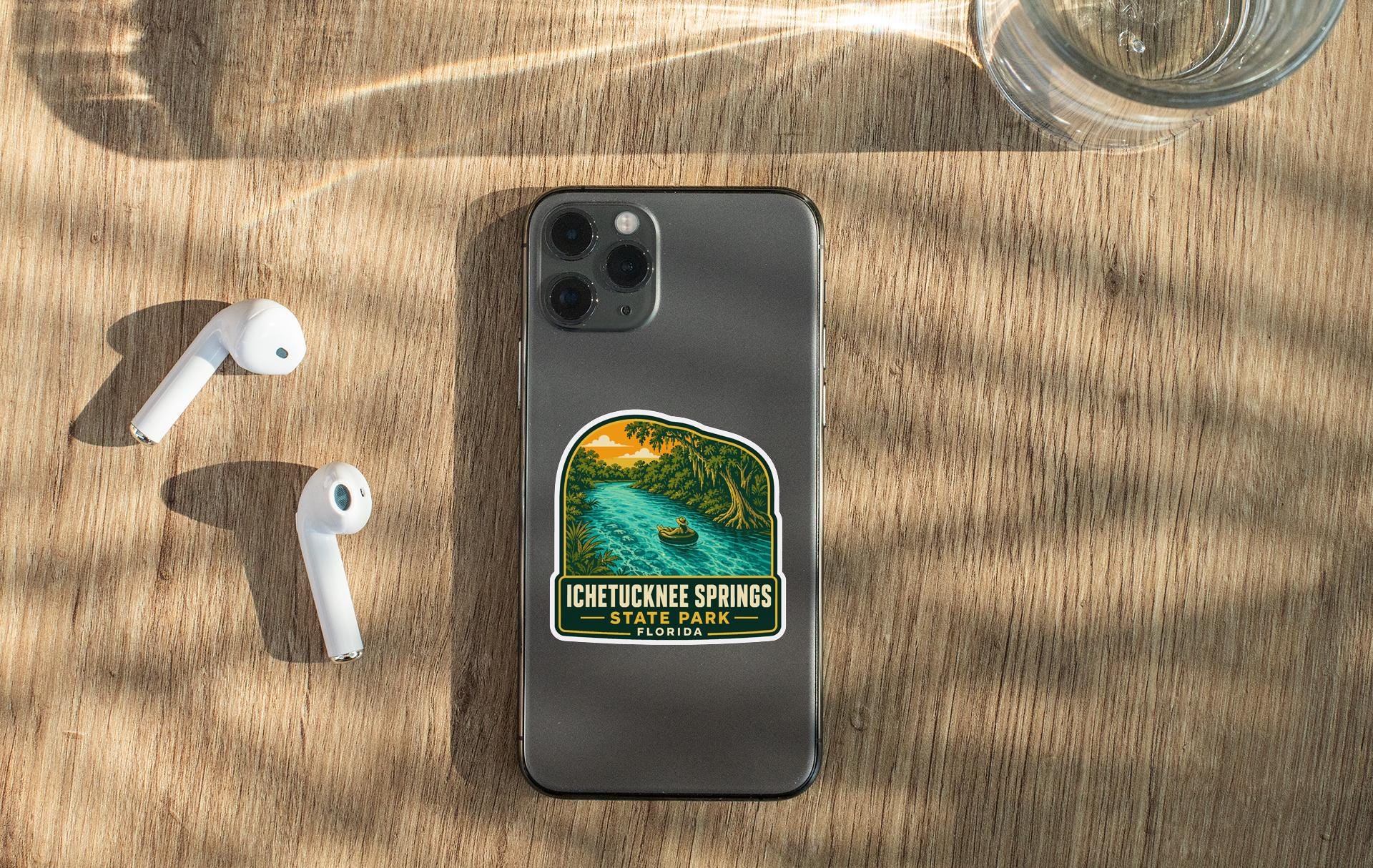 a smartphone with a sticker on it, placed on a wooden surface alongside a pair of white earbuds and a glass of water.