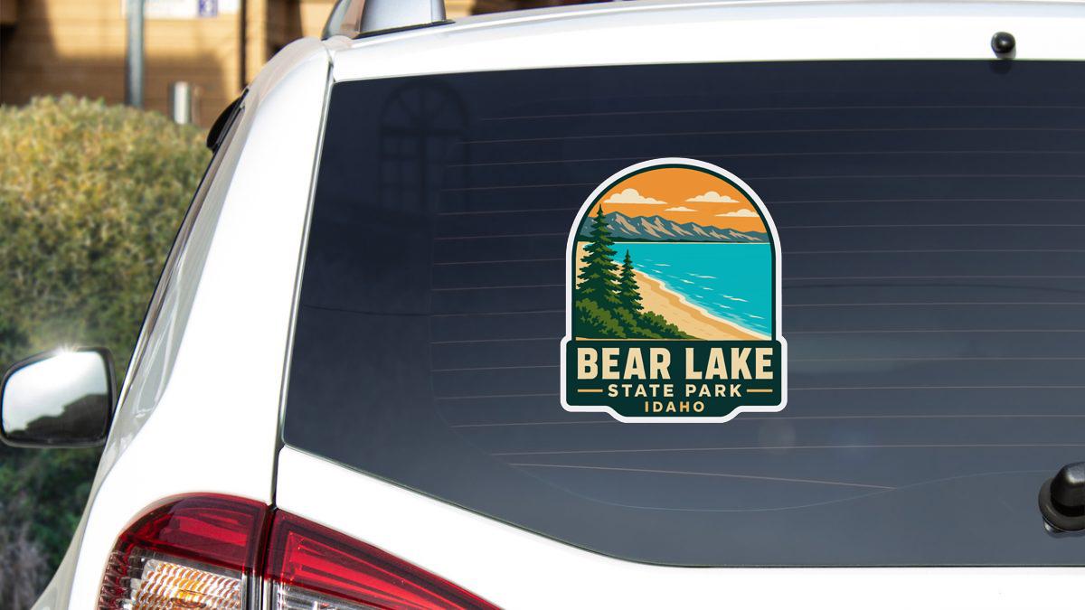 A sticker on the rear window of a white car, depicting a scenic landscape with mountains, a beach, and a lake.