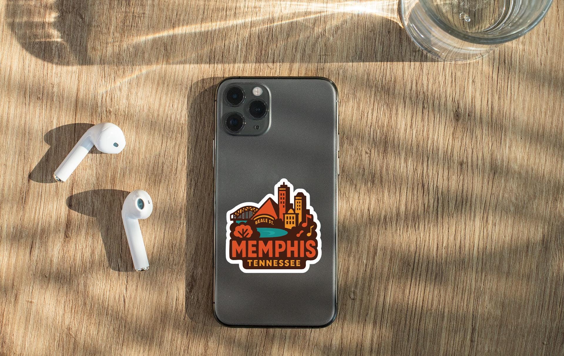 a smartphone with a sticker on the back, placed on a wooden surface alongside a pair of white earbuds and a glass of water.
