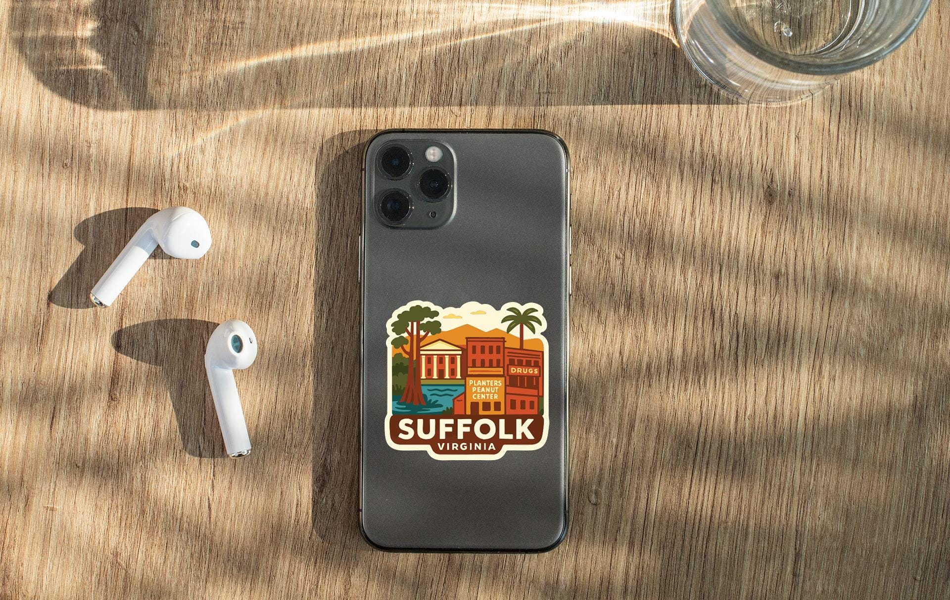 a smartphone with a sticker of a city on it, placed on a wooden surface alongside a pair of white earbuds and a glass of water.