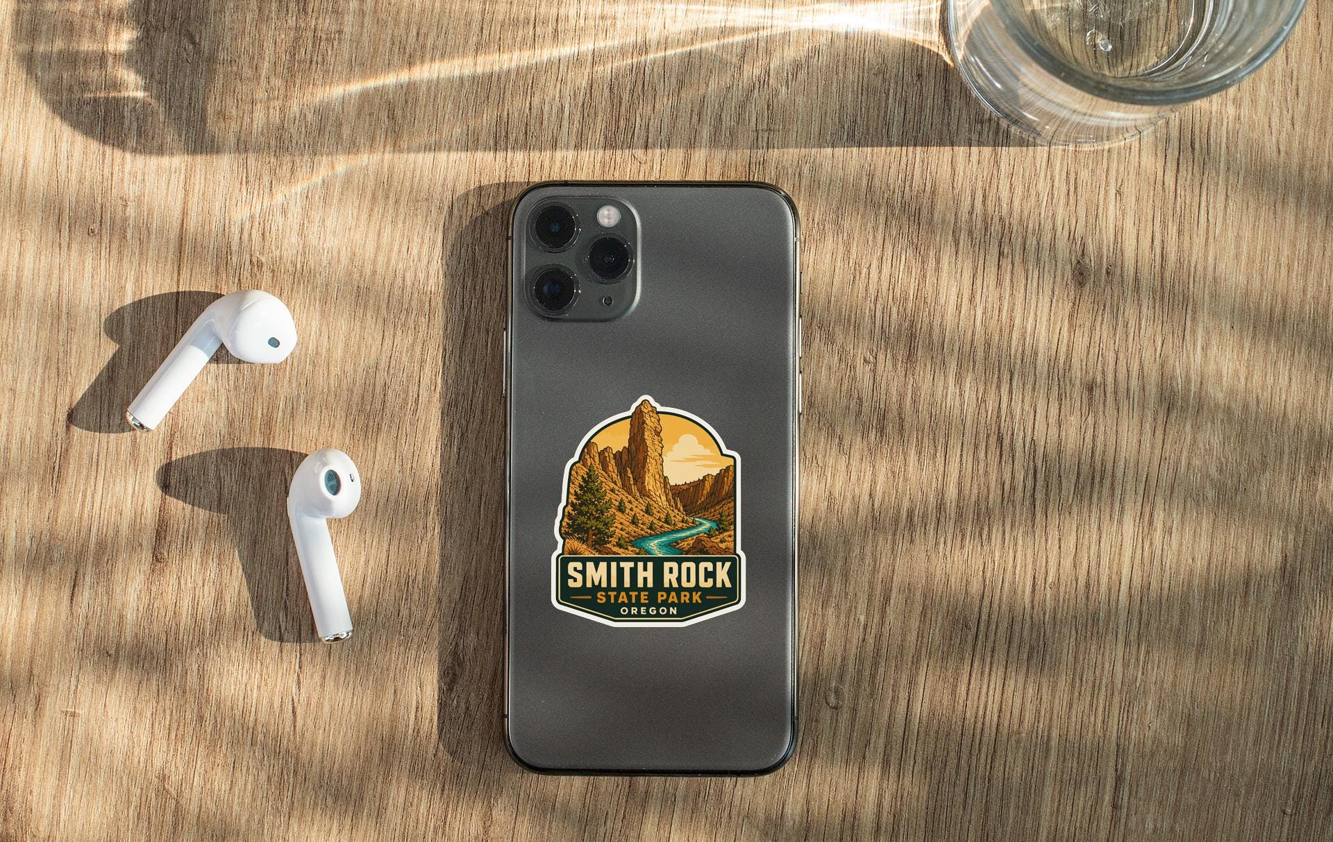 a smartphone with a sticker of Smith Rock State Park on its back, along with a pair of white earbuds and a glass of water on a wooden surface.