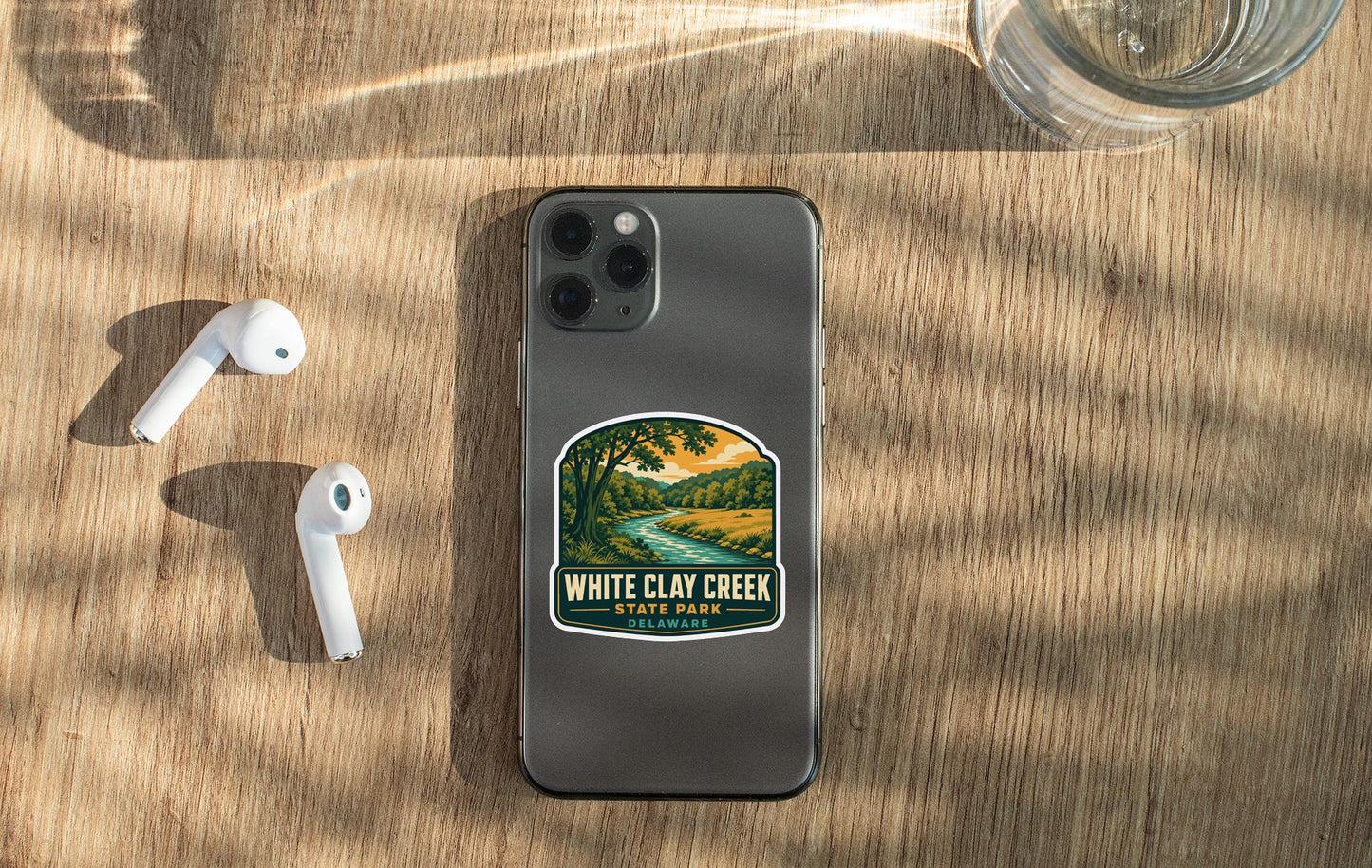 a smartphone with a sticker on the back, along with a pair of white earbuds and a glass of water on a wooden surface.