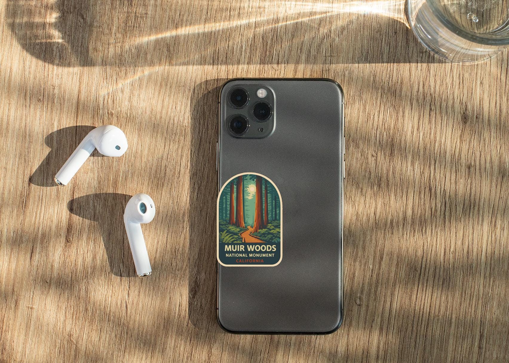 a smartphone with a sticker on the back, placed on a wooden surface alongside a pair of white earbuds and a glass of water.