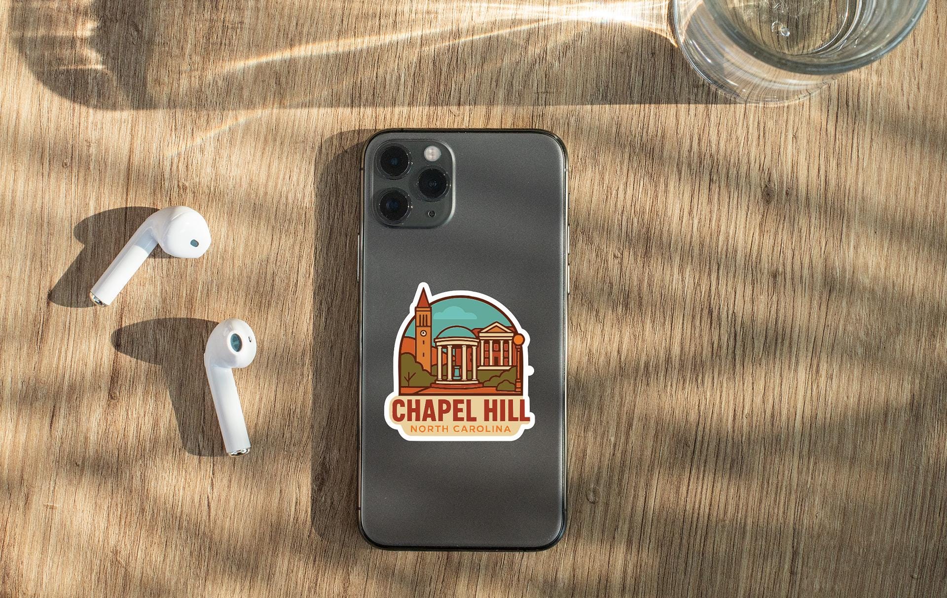 a smartphone with a sticker of Chapel Hill, North Carolina, placed on a wooden surface alongside a pair of white earbuds and a glass of water.