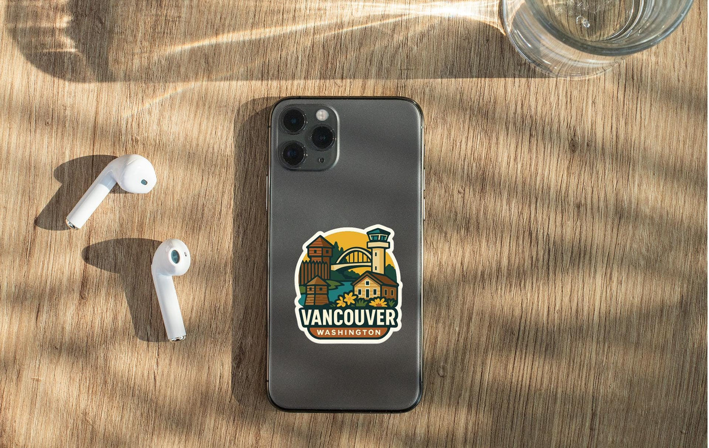 a smartphone with a sticker of Vancouver, Washington, placed on a wooden surface alongside a pair of white earbuds and a glass of water.