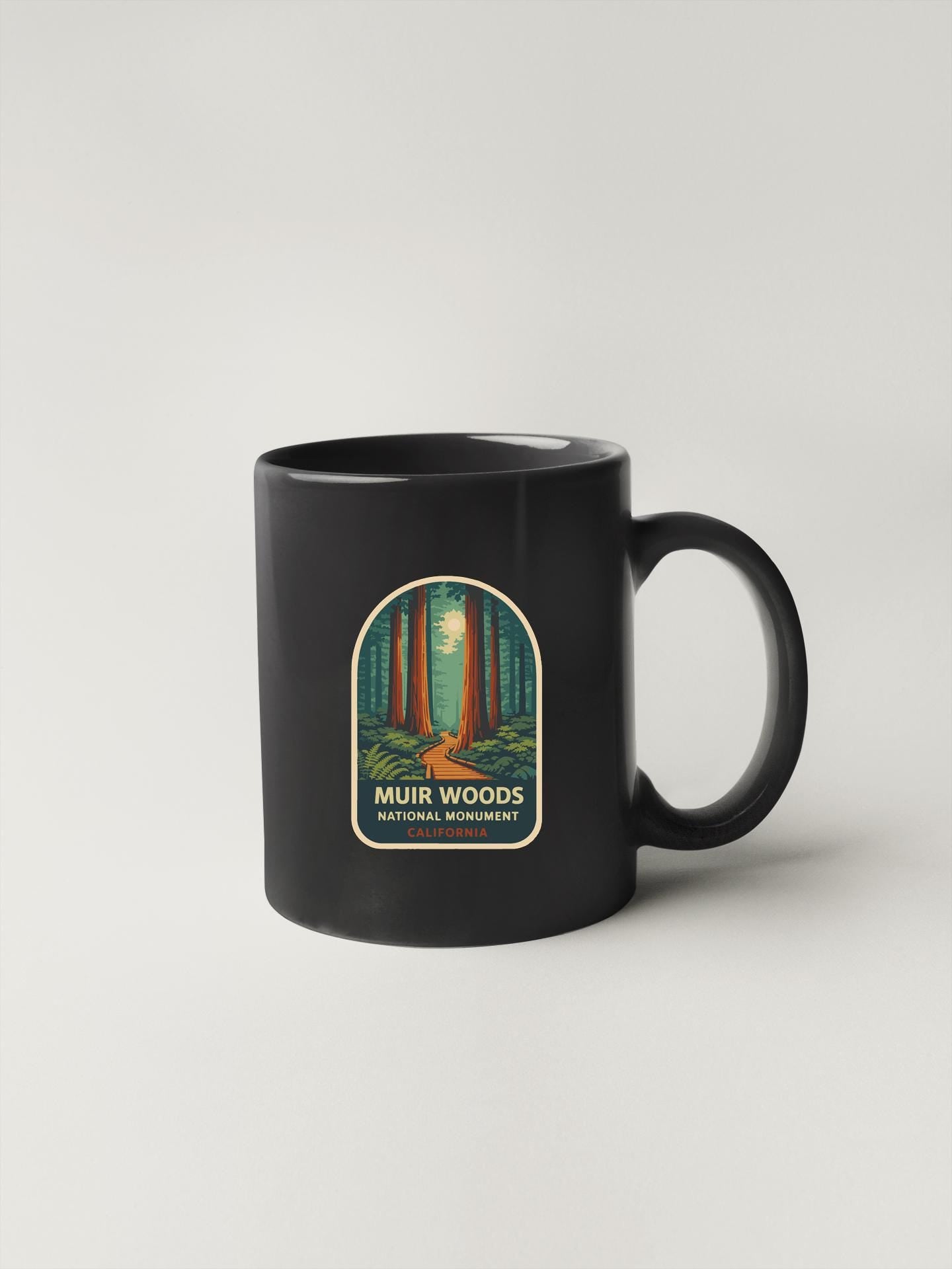 A black coffee mug with a label that says "Muiir Woods National Monument" and features an illustration of a forest scene.