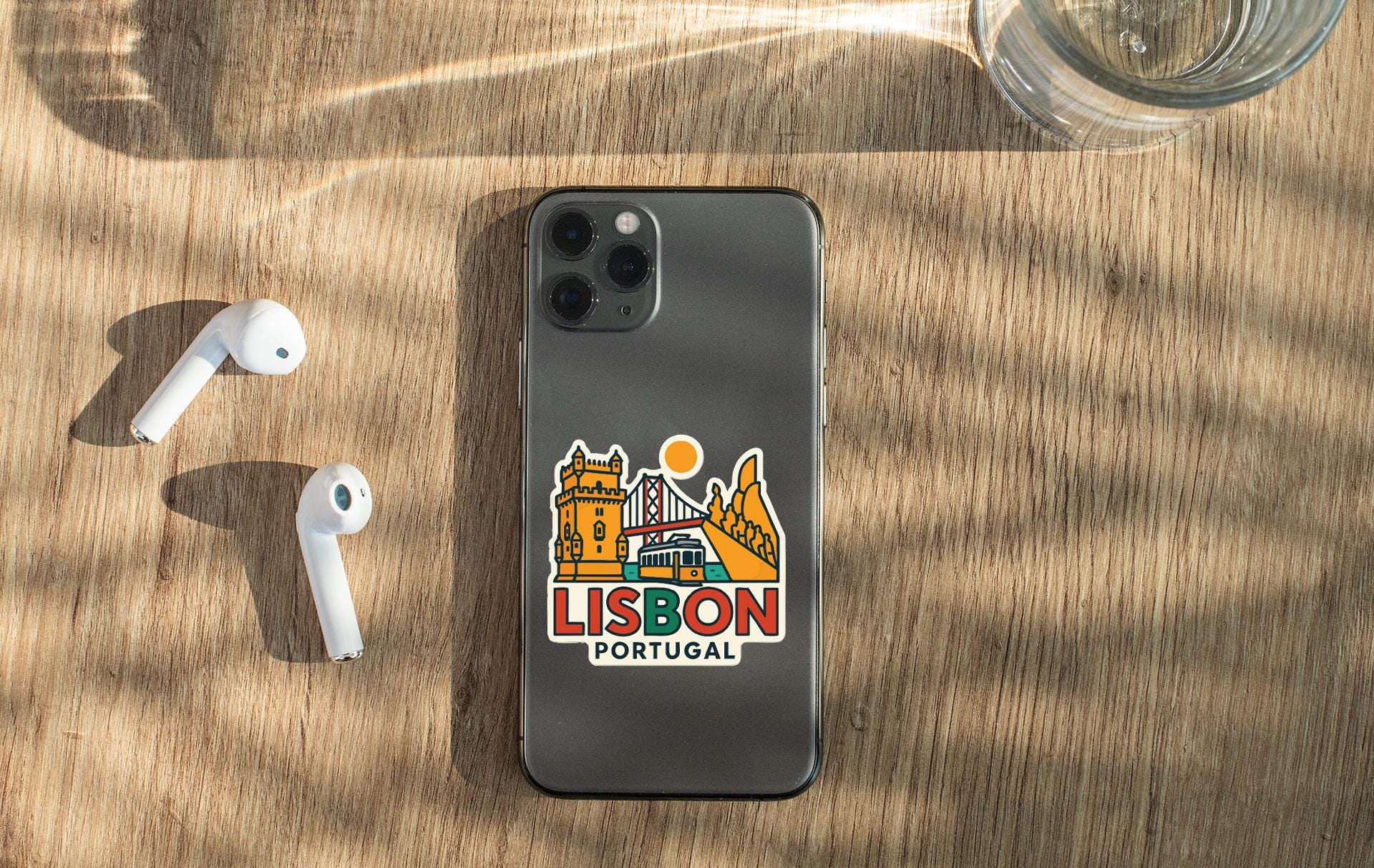 a smartphone with a sticker of Lisbon, Portugal, placed on a wooden surface alongside a pair of white earbuds and a glass of water.