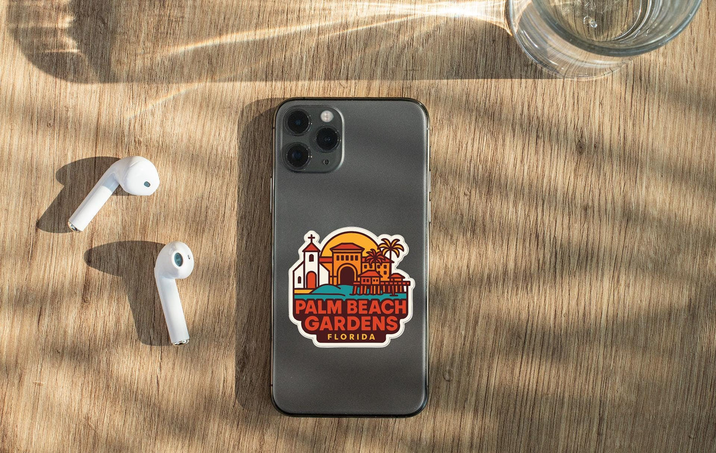 a smartphone with a sticker on the back, placed on a wooden surface alongside a pair of white earbuds and a glass of water.