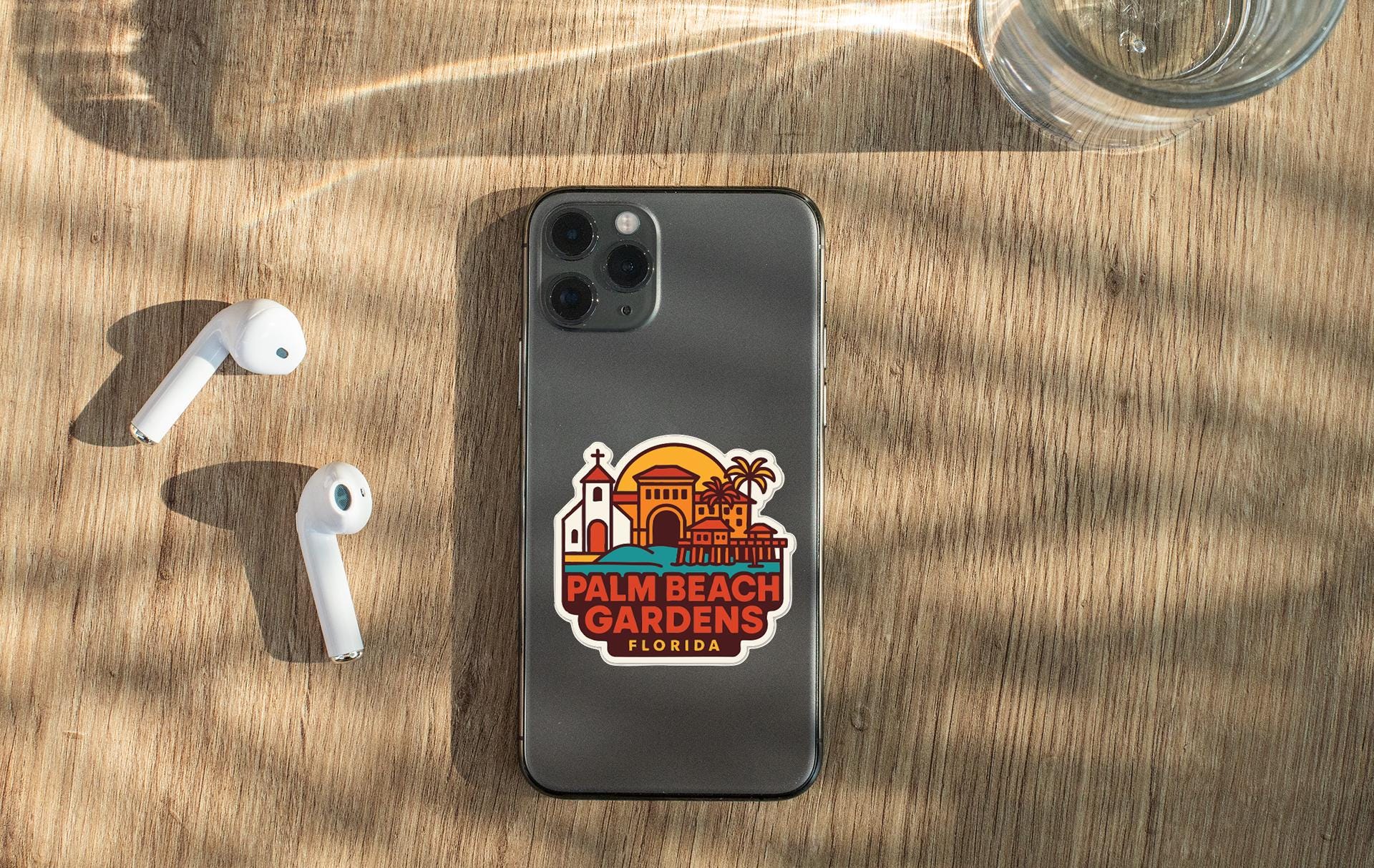 a smartphone with a sticker on the back, placed on a wooden surface alongside a pair of white earbuds and a glass of water.