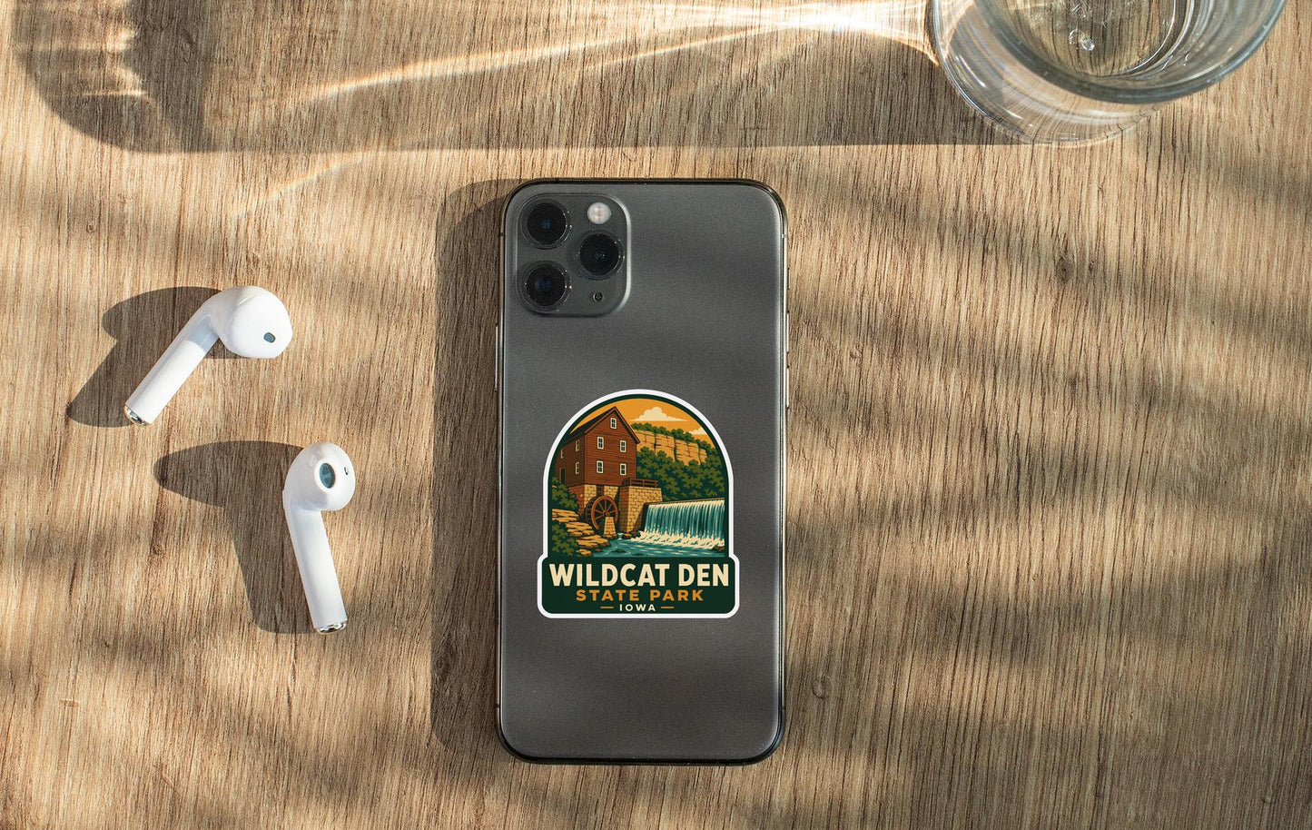 a smartphone with a sticker on it, placed on a wooden surface alongside a pair of white earbuds and a glass of water.