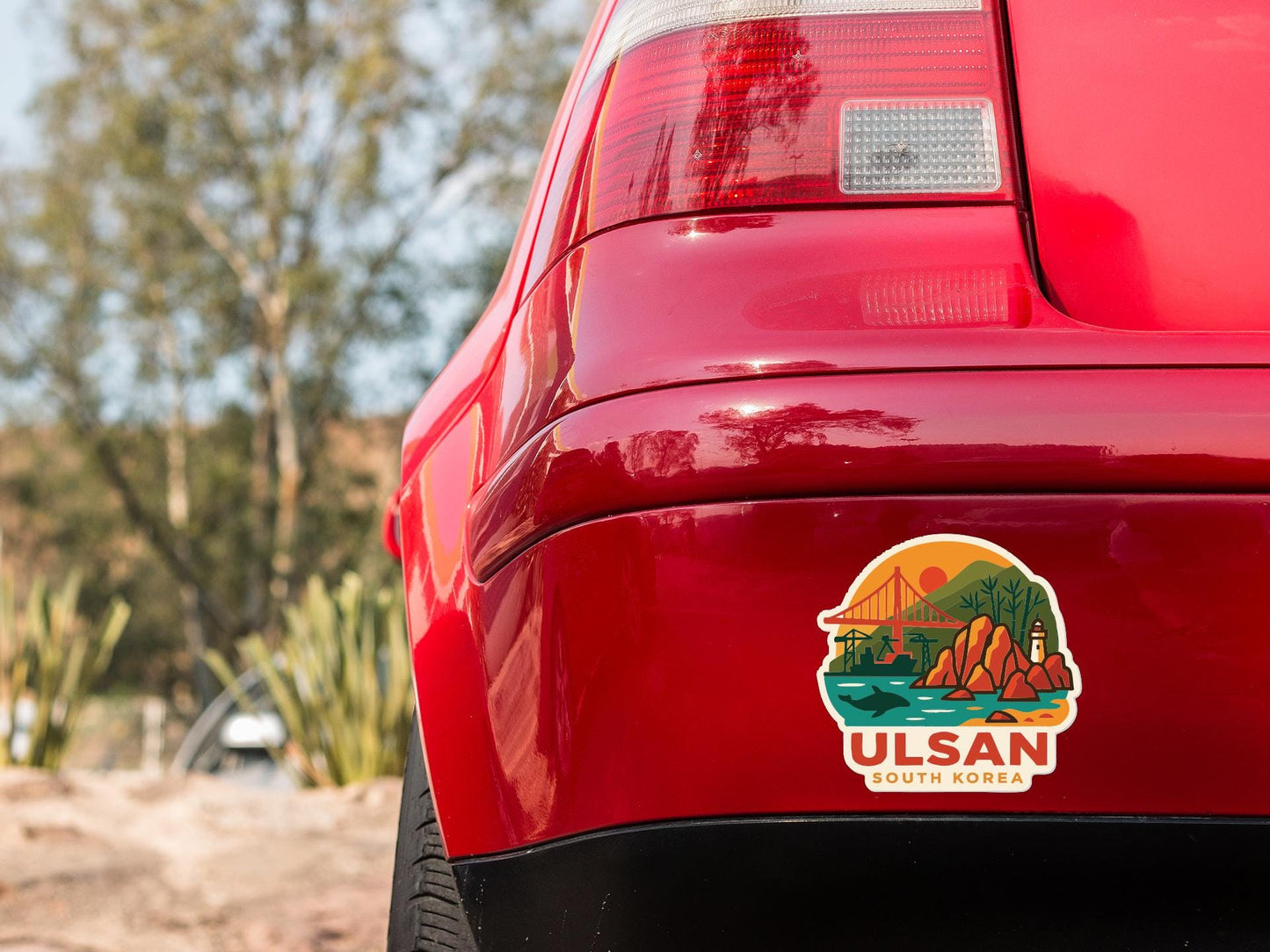 the rear end of a red car with a sticker on it, featuring a scenic landscape with mountains, a bridge, and water.