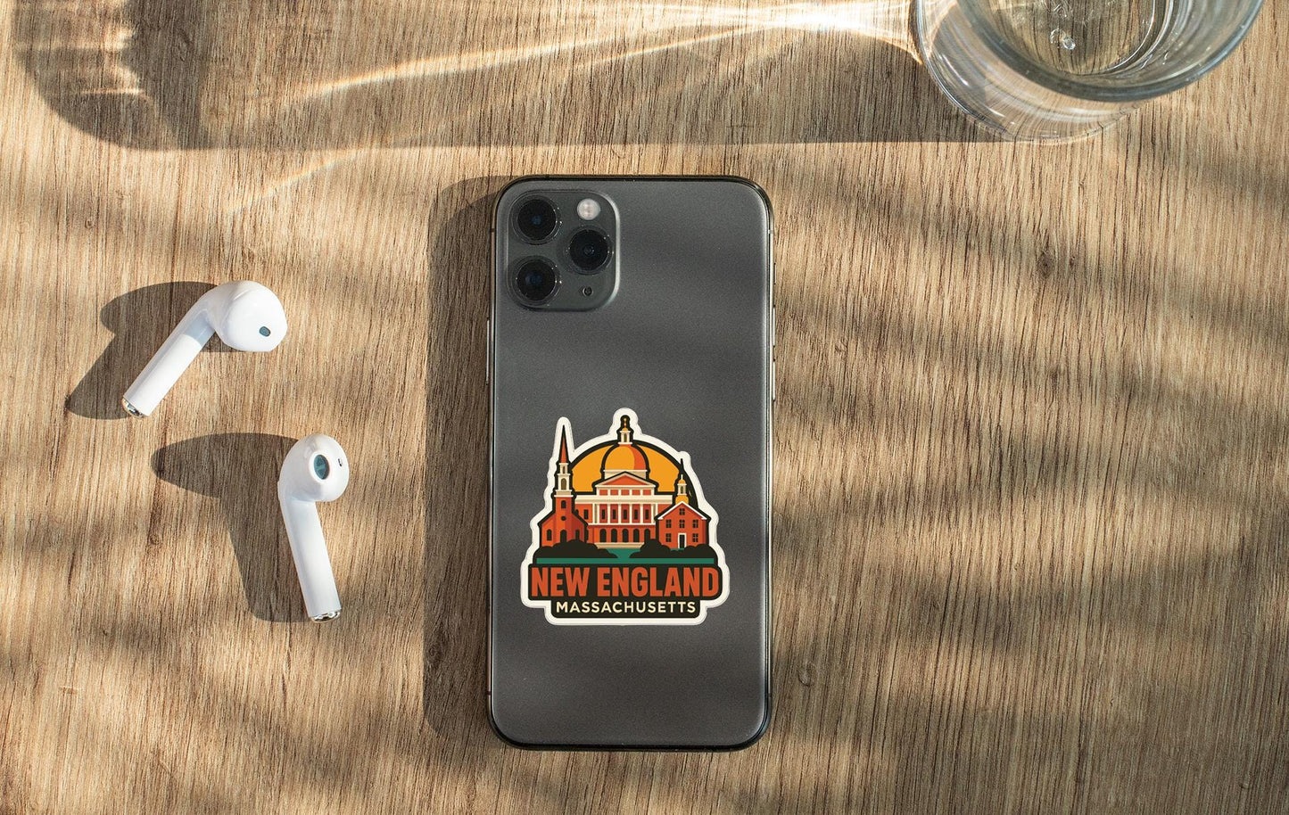 a smartphone with a sticker of the New England state of Massachusetts, along with a pair of white earbuds and a glass of water on a wooden surface.