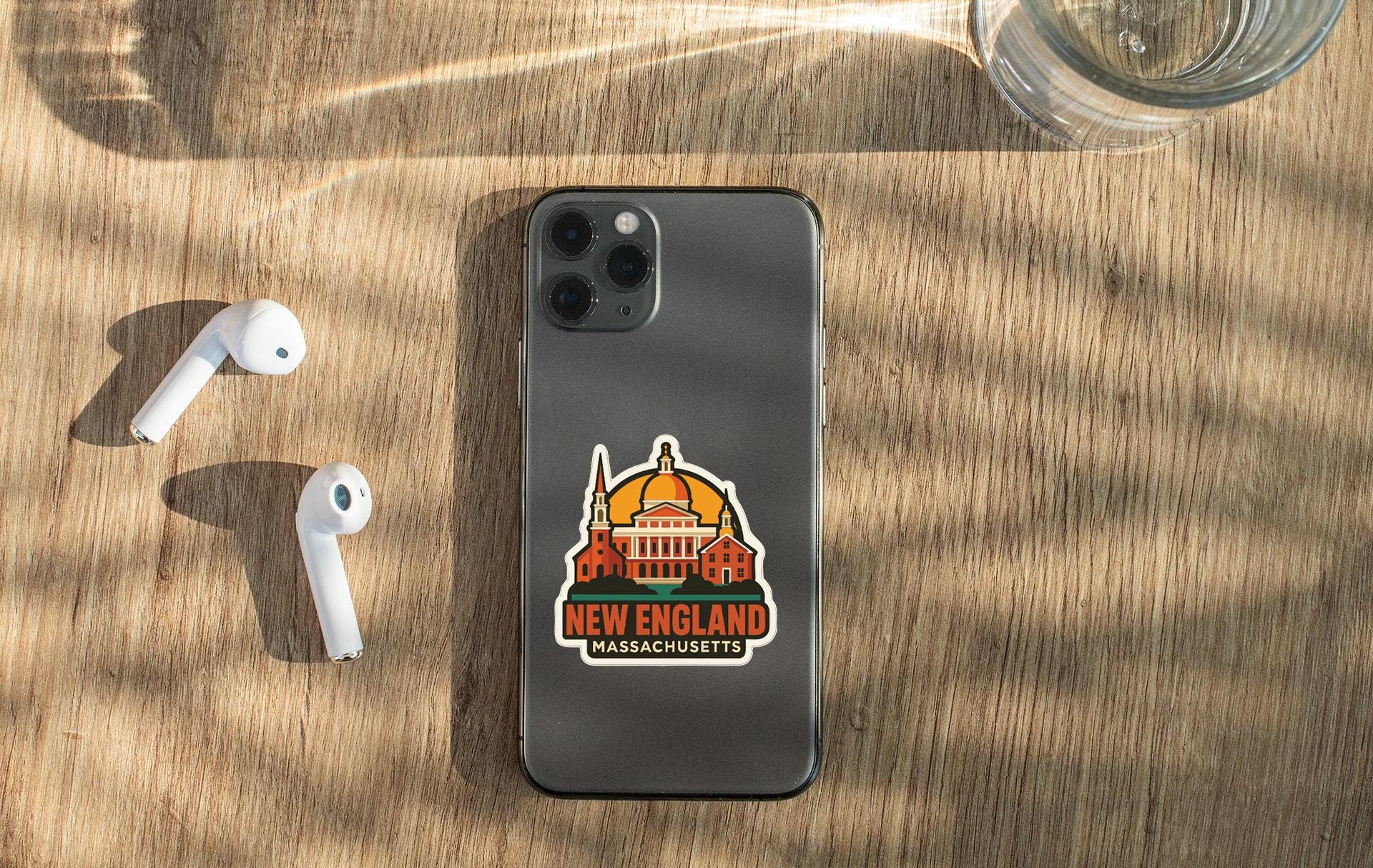 a smartphone with a sticker of the New England state of Massachusetts, along with a pair of white earbuds and a glass of water on a wooden surface.