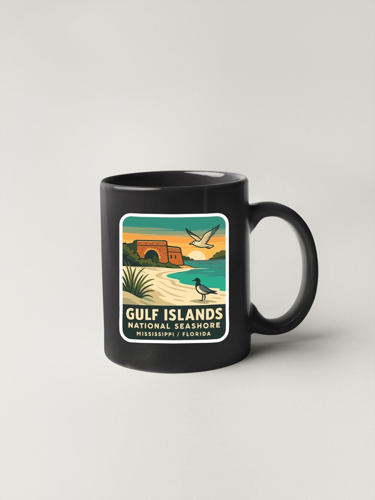 A black coffee mug with a graphic design depicting a beach scene and a bird flying over the water.
