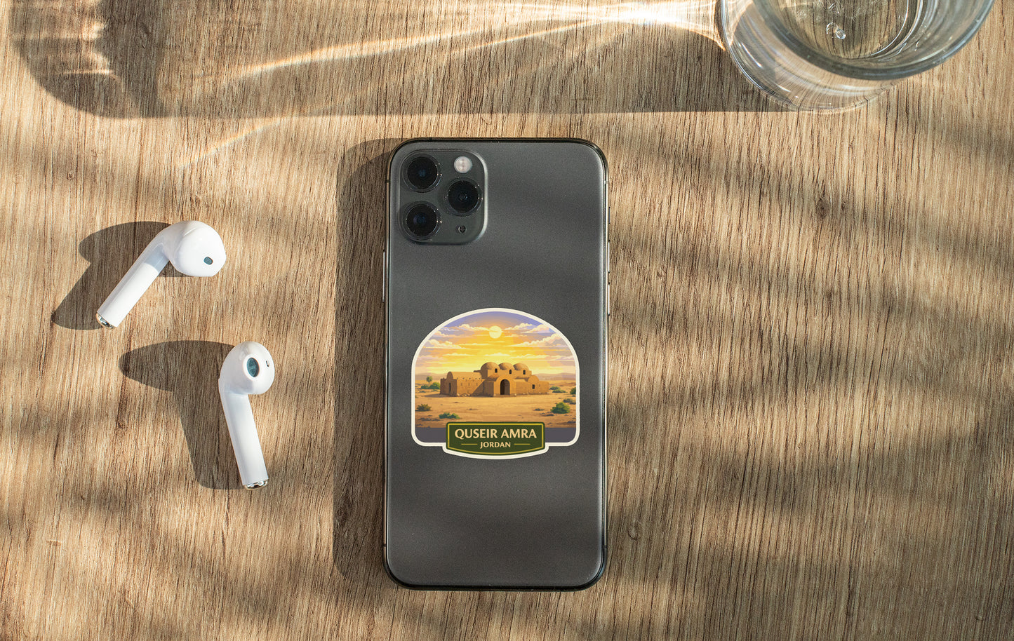a smartphone with a sticker of Quesker Amara, a desert town in Saudi Arabia, placed on a wooden surface alongside a pair of white earbuds and a glass of water.