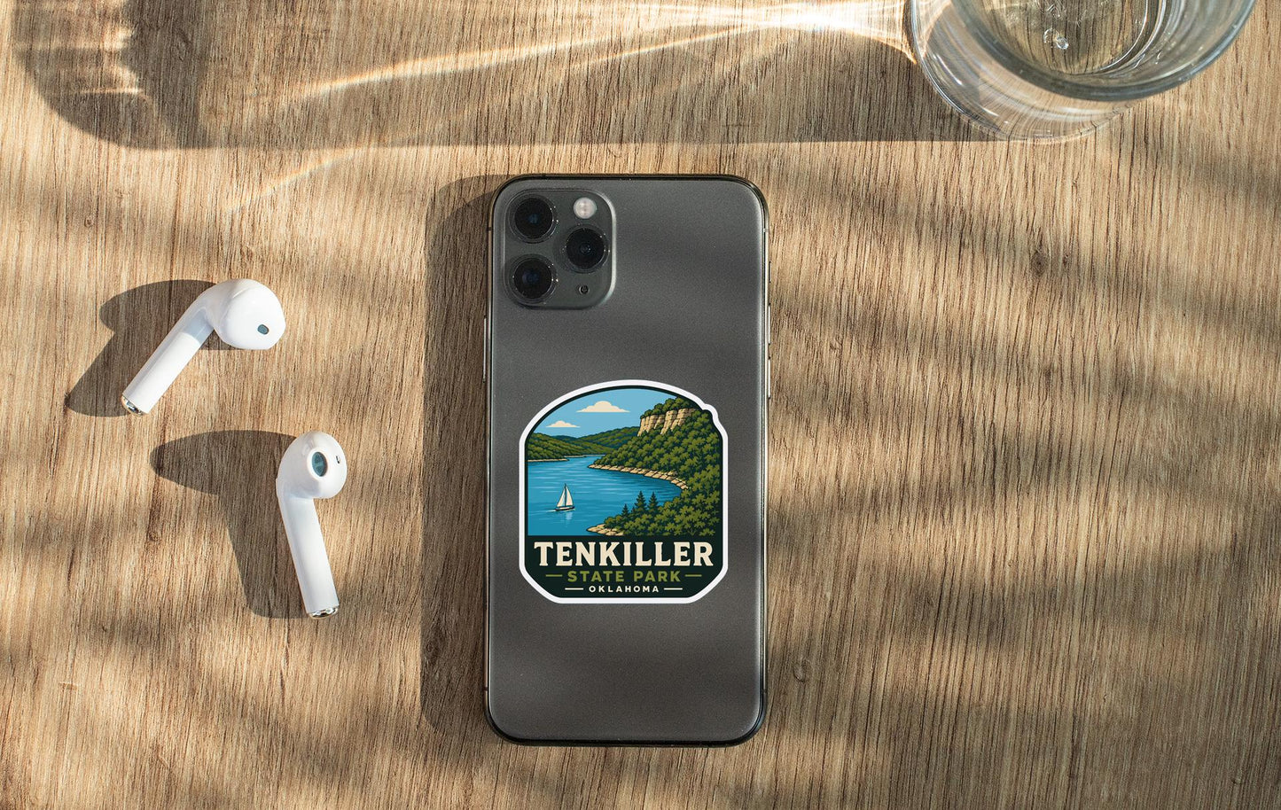 a smartphone with a sticker of Ten Killer State Park on it, placed on a wooden surface alongside a pair of white earbuds and a glass of water.