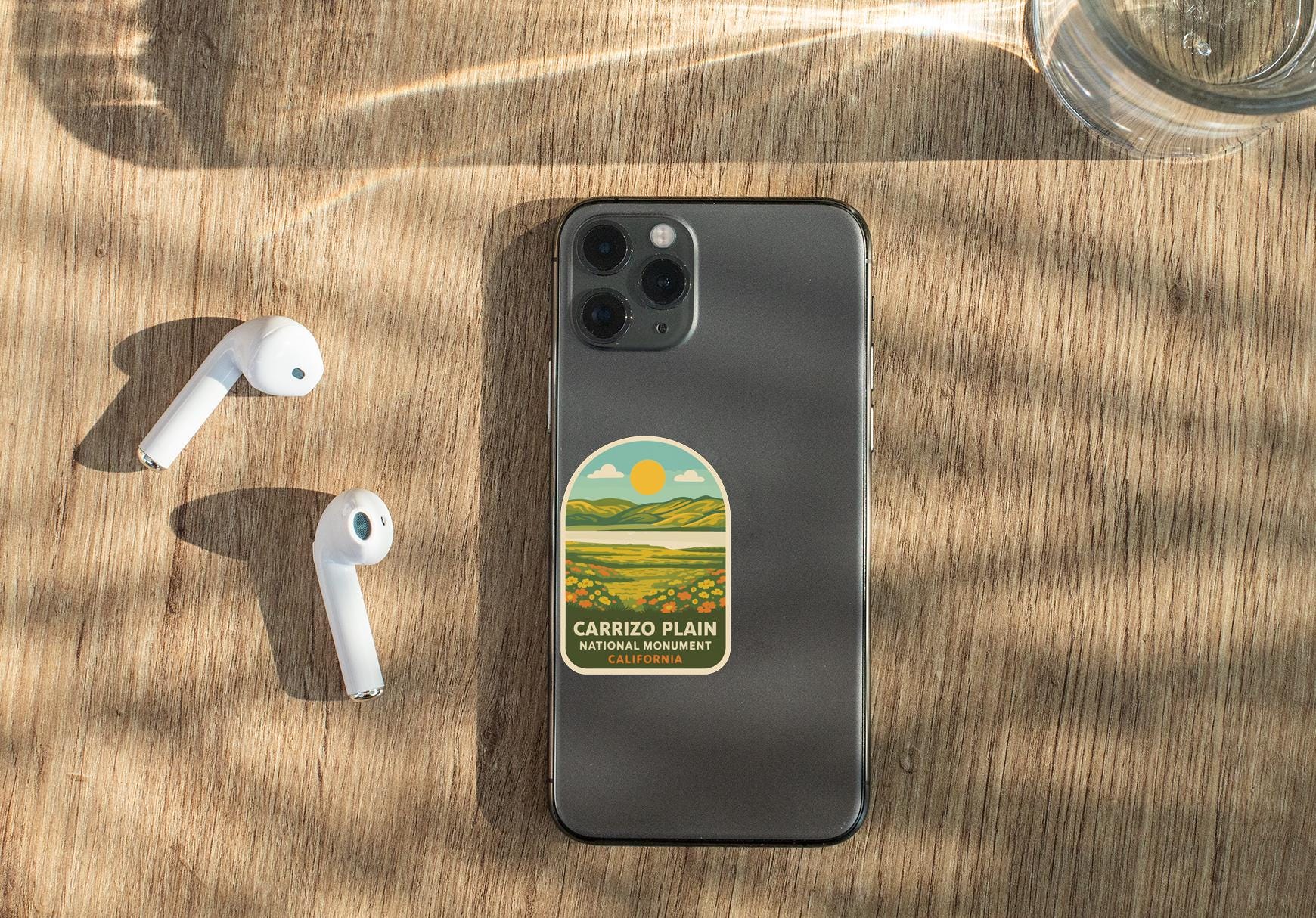 a smartphone with a sticker on it, placed on a wooden surface alongside a pair of white earbuds and a glass of water.