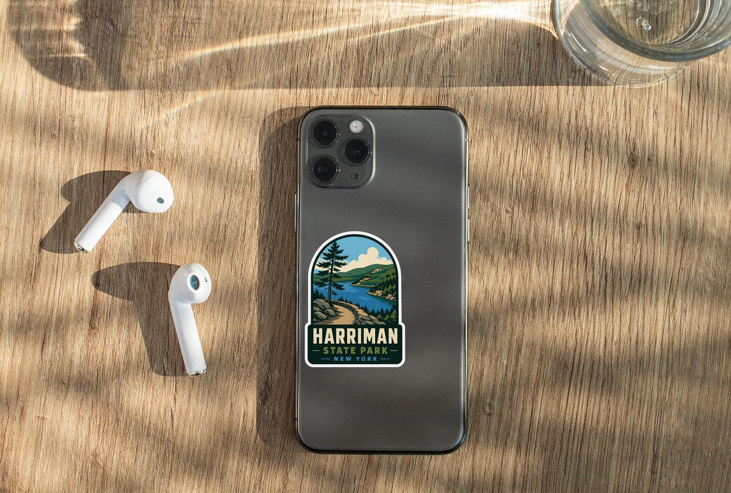 a smartphone with a sticker on the back, placed on a wooden surface alongside a pair of white earbuds and a glass of water.