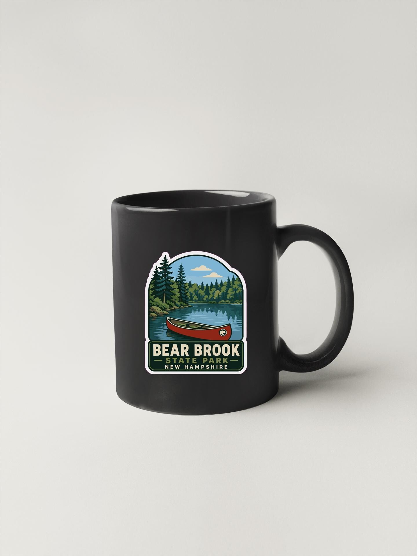 A black coffee mug with a sticker depicting a canoe on a lake surrounded by trees, with the text "BEAR BROOK NEW HAMPSHIRE" printed on it.