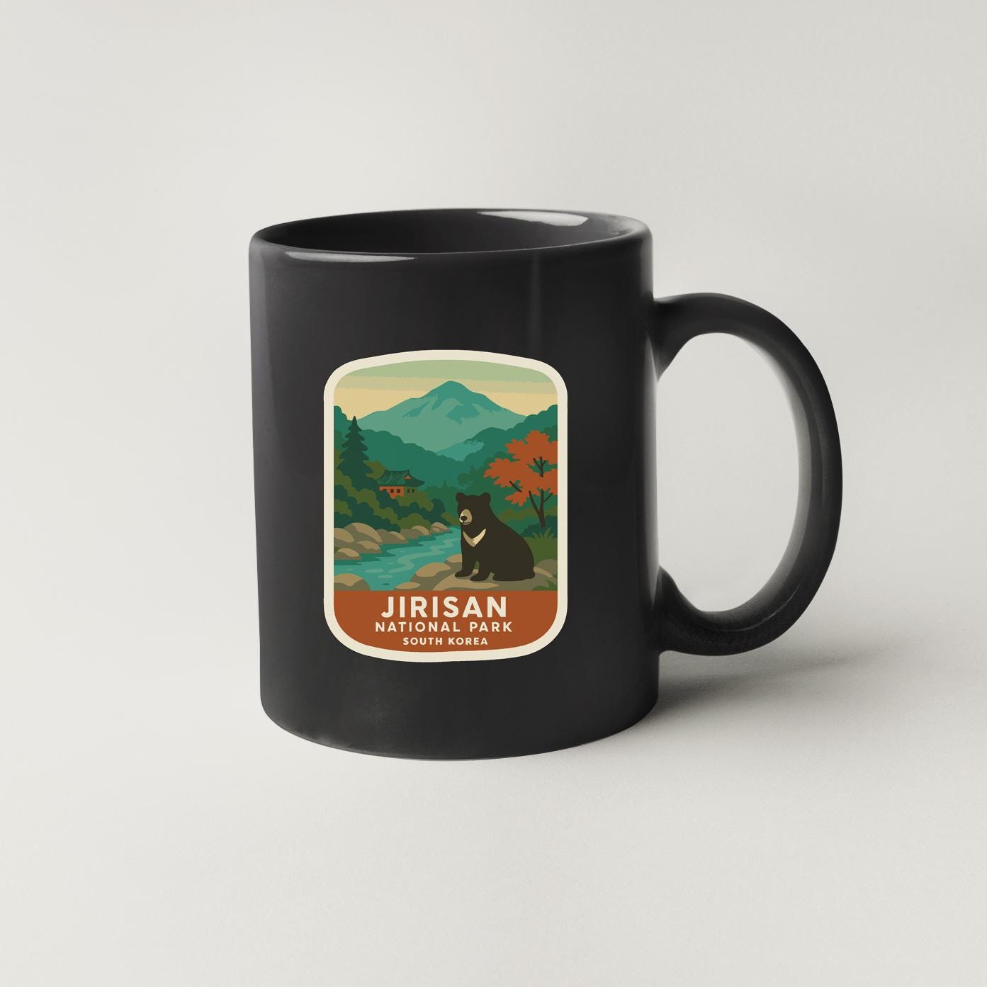 A black coffee mug with a label that features an illustration of a bear in a scenic landscape, along with the text "Jirisan National Park SOUTH KOREA".