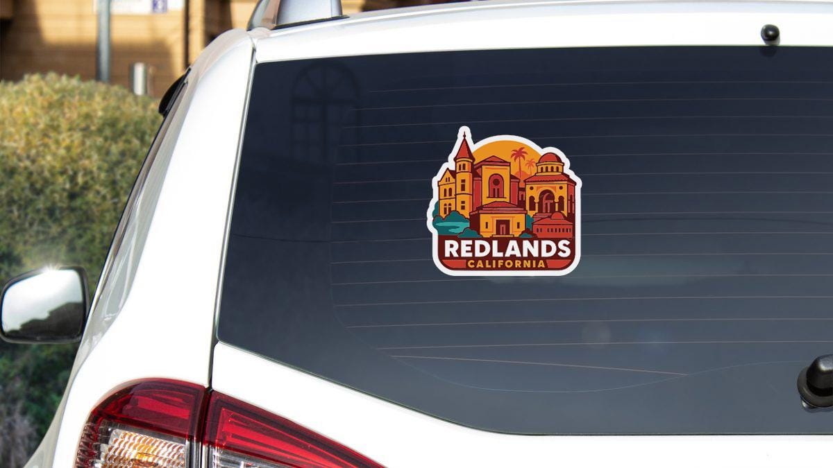A sticker on the rear window of a white car, depicting a cityscape with buildings and a palm tree, representing the city of Redlands, California.
