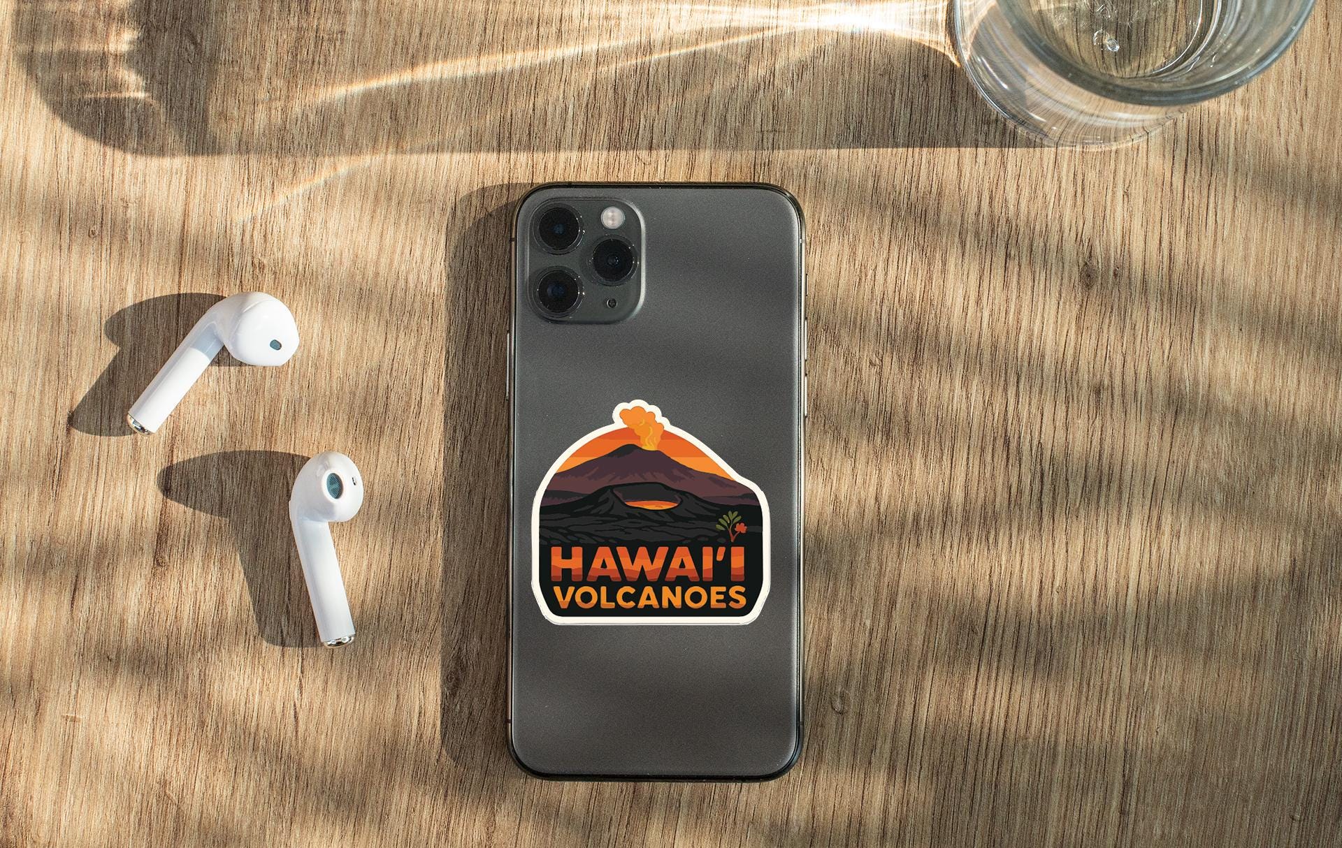 a smartphone with a sticker on the back, placed on a wooden surface alongside a pair of white earbuds and a glass of water.