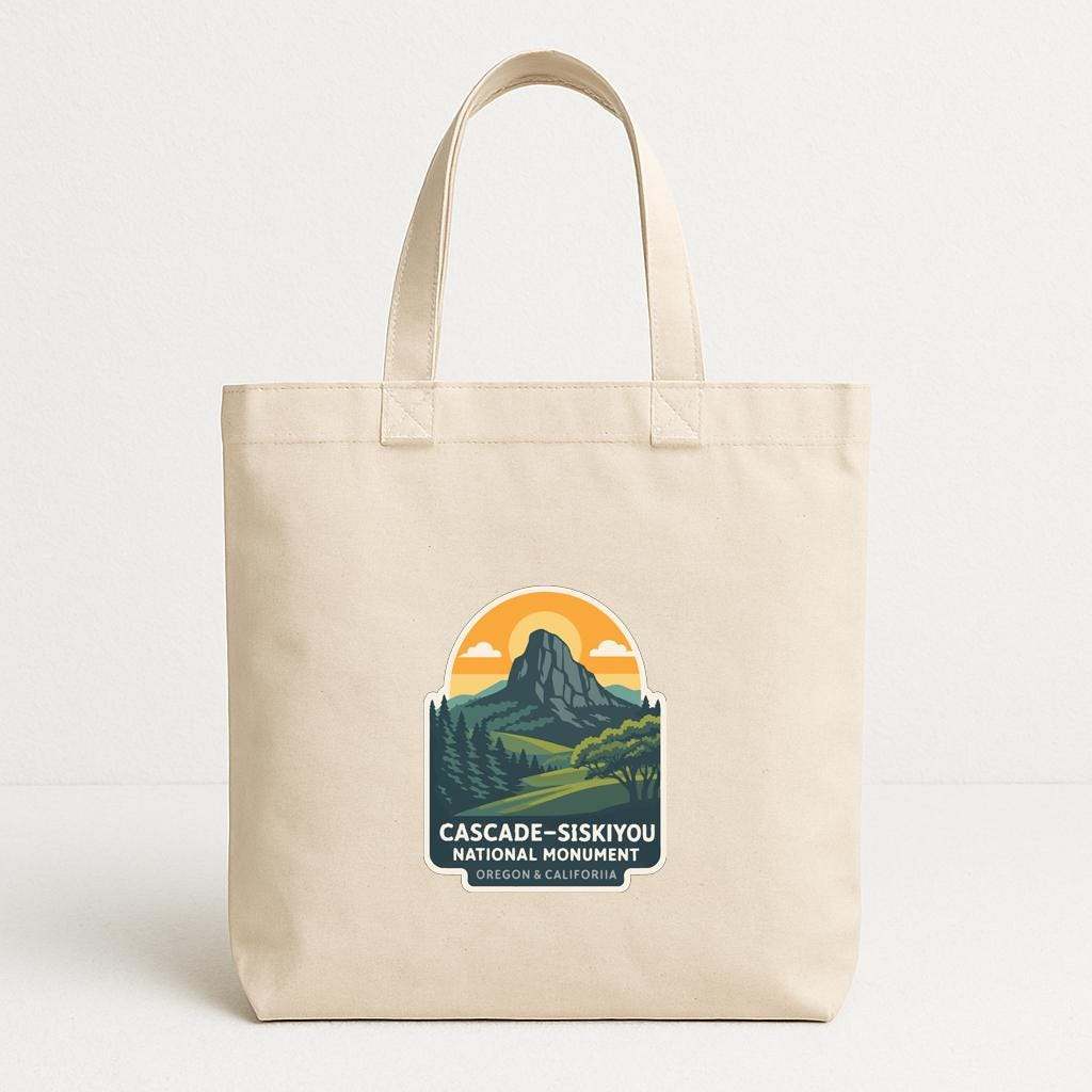 A beige tote bag with a printed design featuring a mountain landscape and the text "CASCADE-SISKYOU NATIONAL MONUMENT" printed on it.