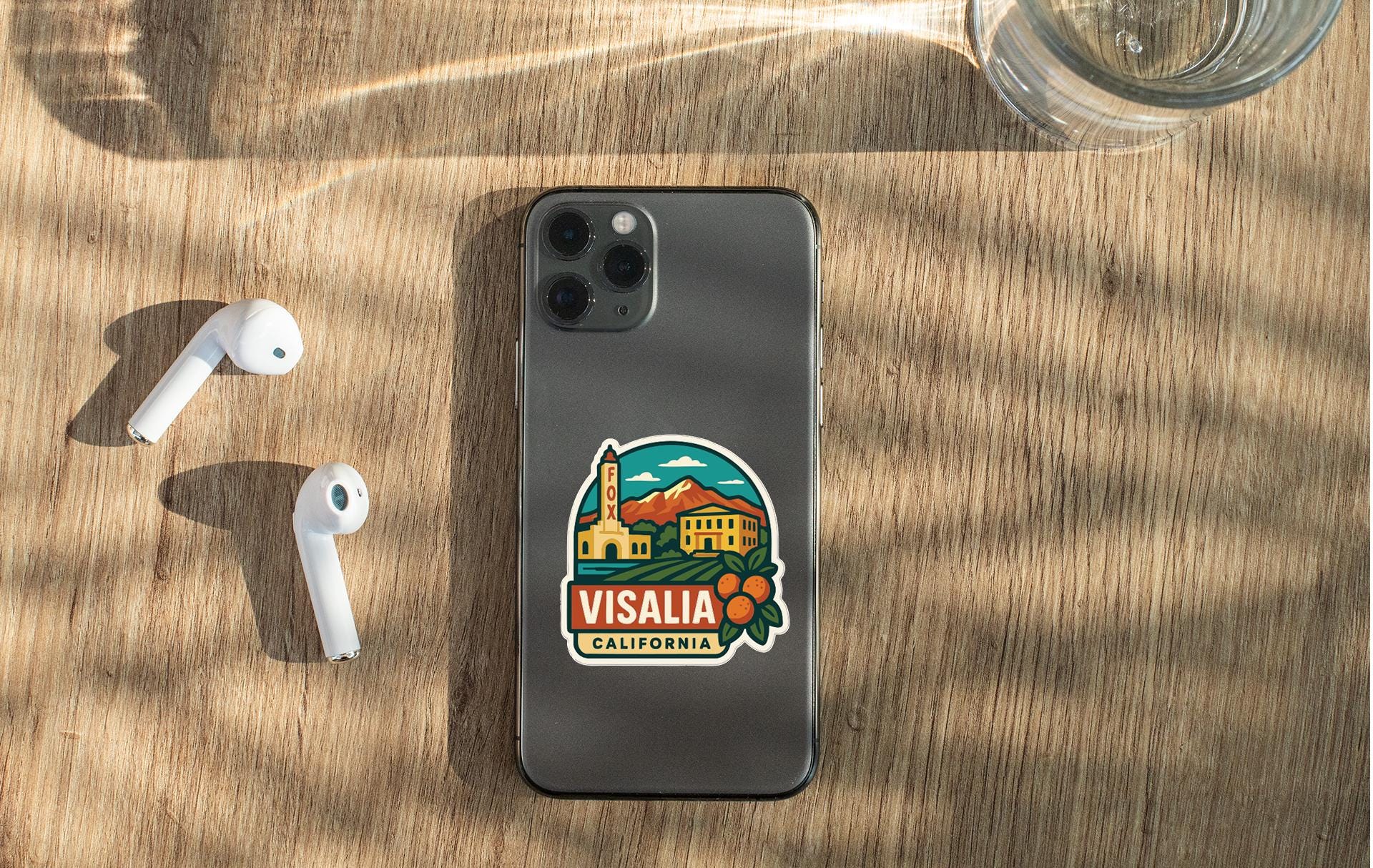 a smartphone with a sticker of a scenic California town, along with a pair of white earbuds and a glass of water on a wooden surface.