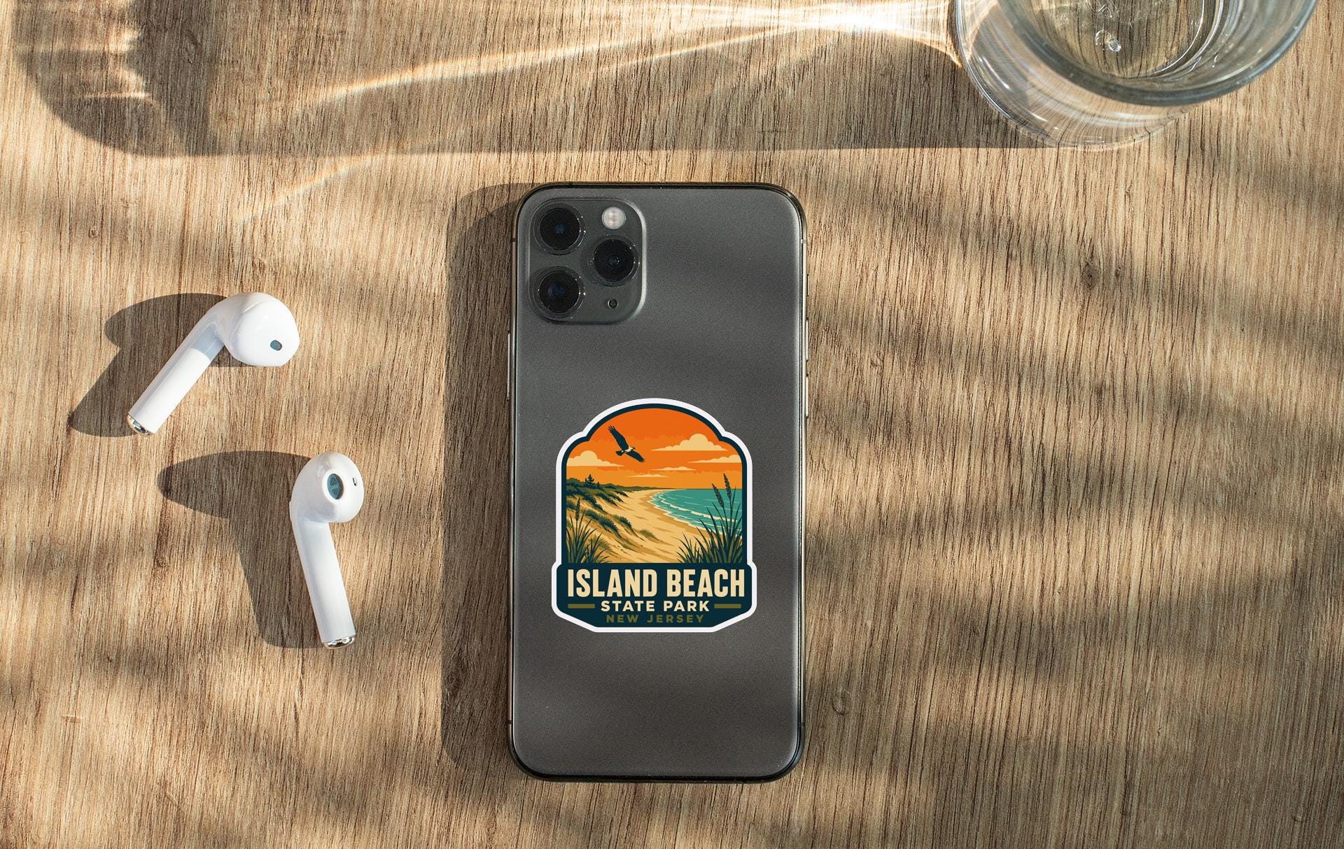 a smartphone with a sticker on the back, placed on a wooden surface alongside a pair of white earbuds and a glass of water.