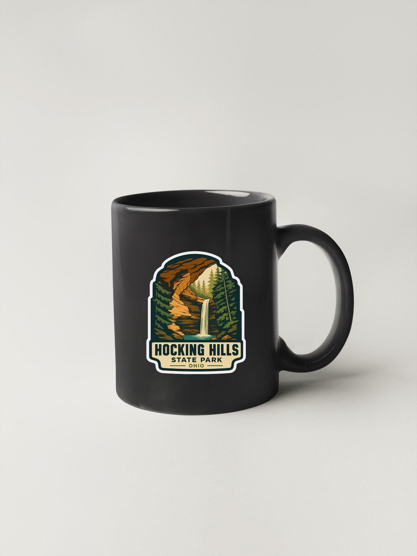 A black coffee mug with a graphic design depicting a scenic landscape, including a waterfall and pine trees, along with the text "HOCKING HILLS STATE PARK".