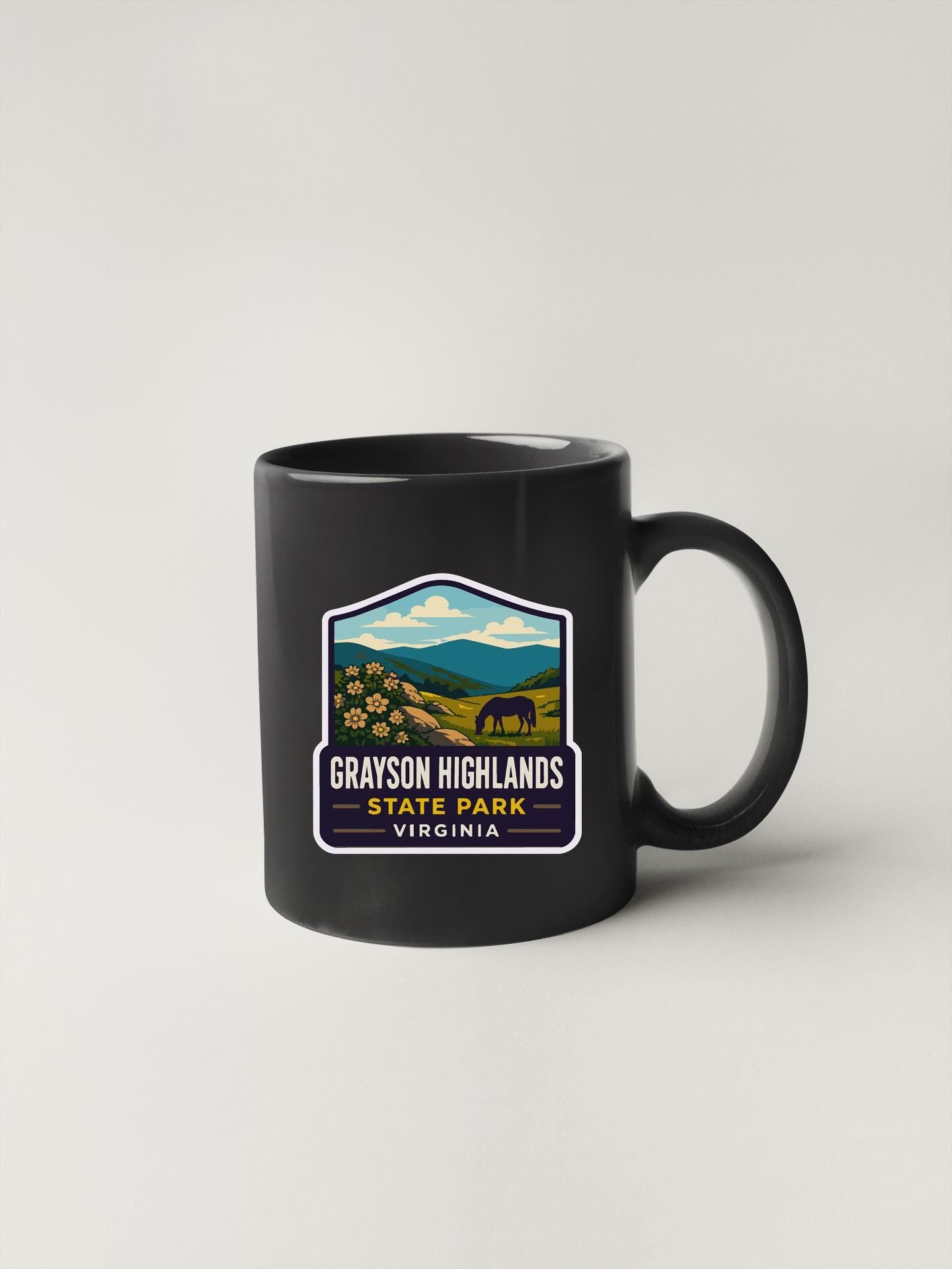 A black coffee mug with a graphic design featuring a scenic landscape with mountains, a horse, and flowers.