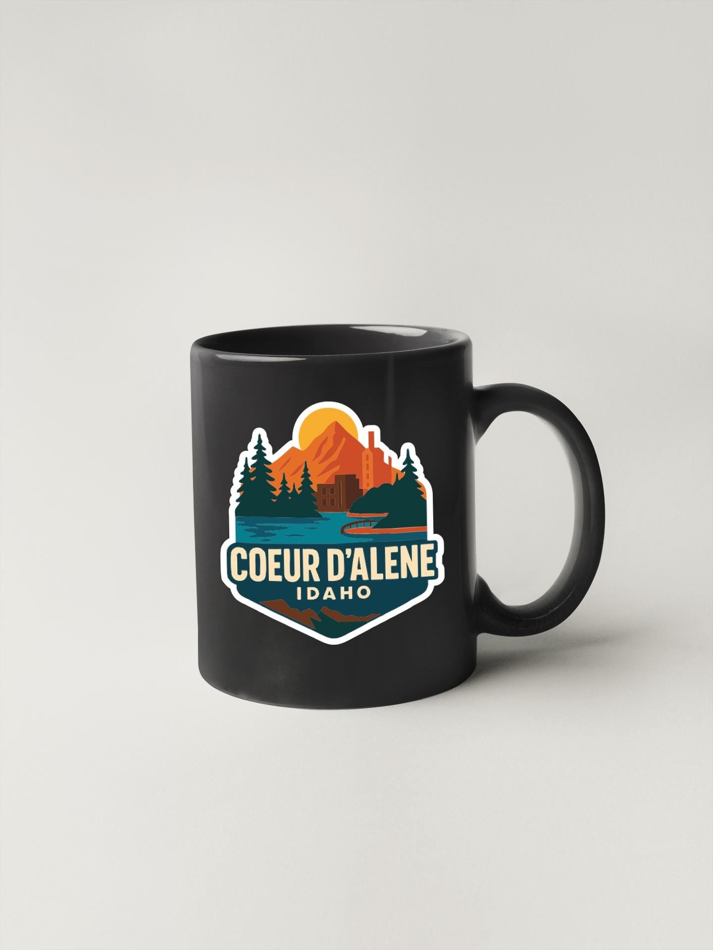 A black coffee mug with a colorful graphic design featuring a mountain landscape and the text "COEUR D'ALENE IDAHO".