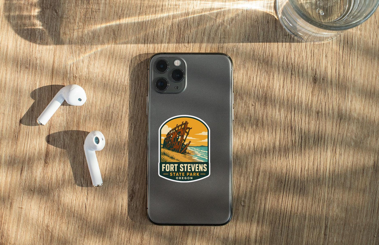 a smartphone with a sticker on the back, placed on a wooden surface alongside a pair of white earbuds and a glass of water.