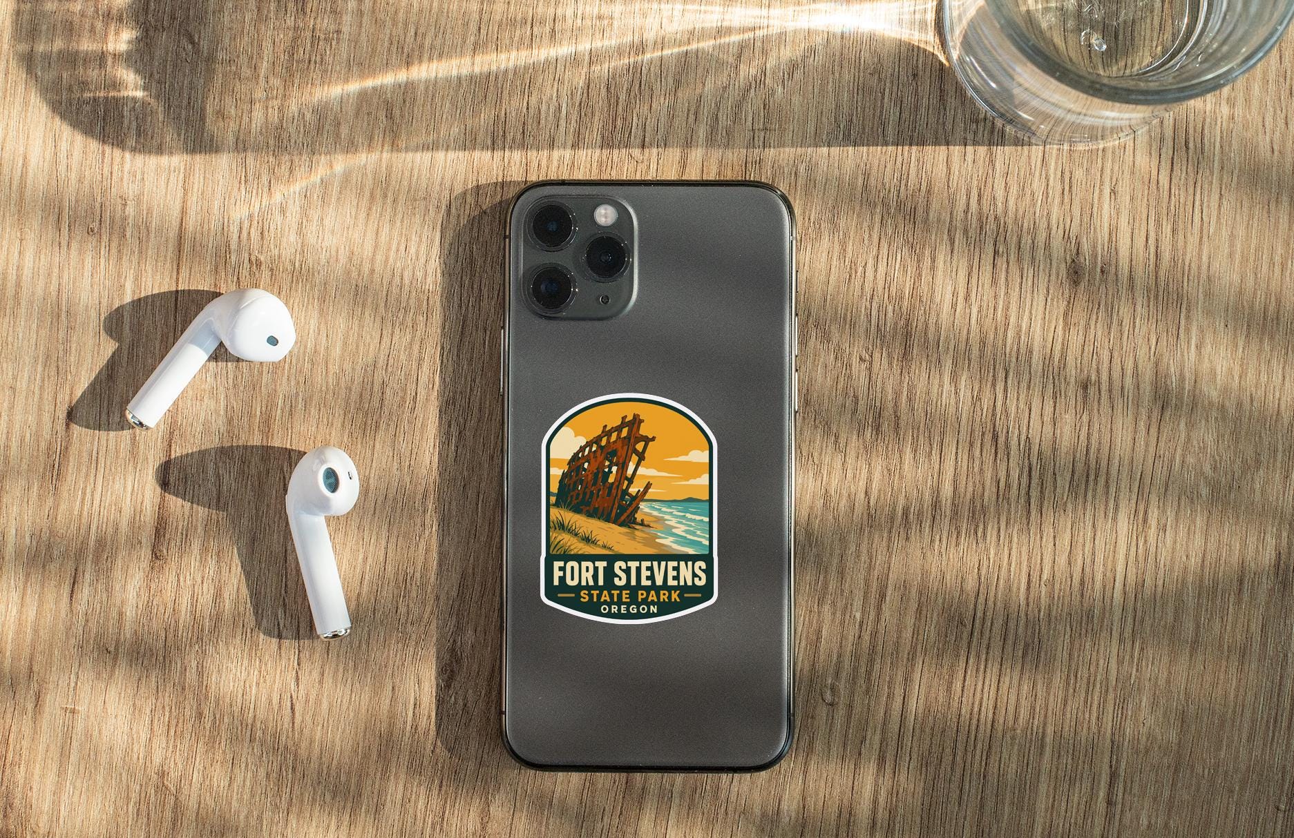 a smartphone with a sticker on the back, placed on a wooden surface alongside a pair of white earbuds and a glass of water.