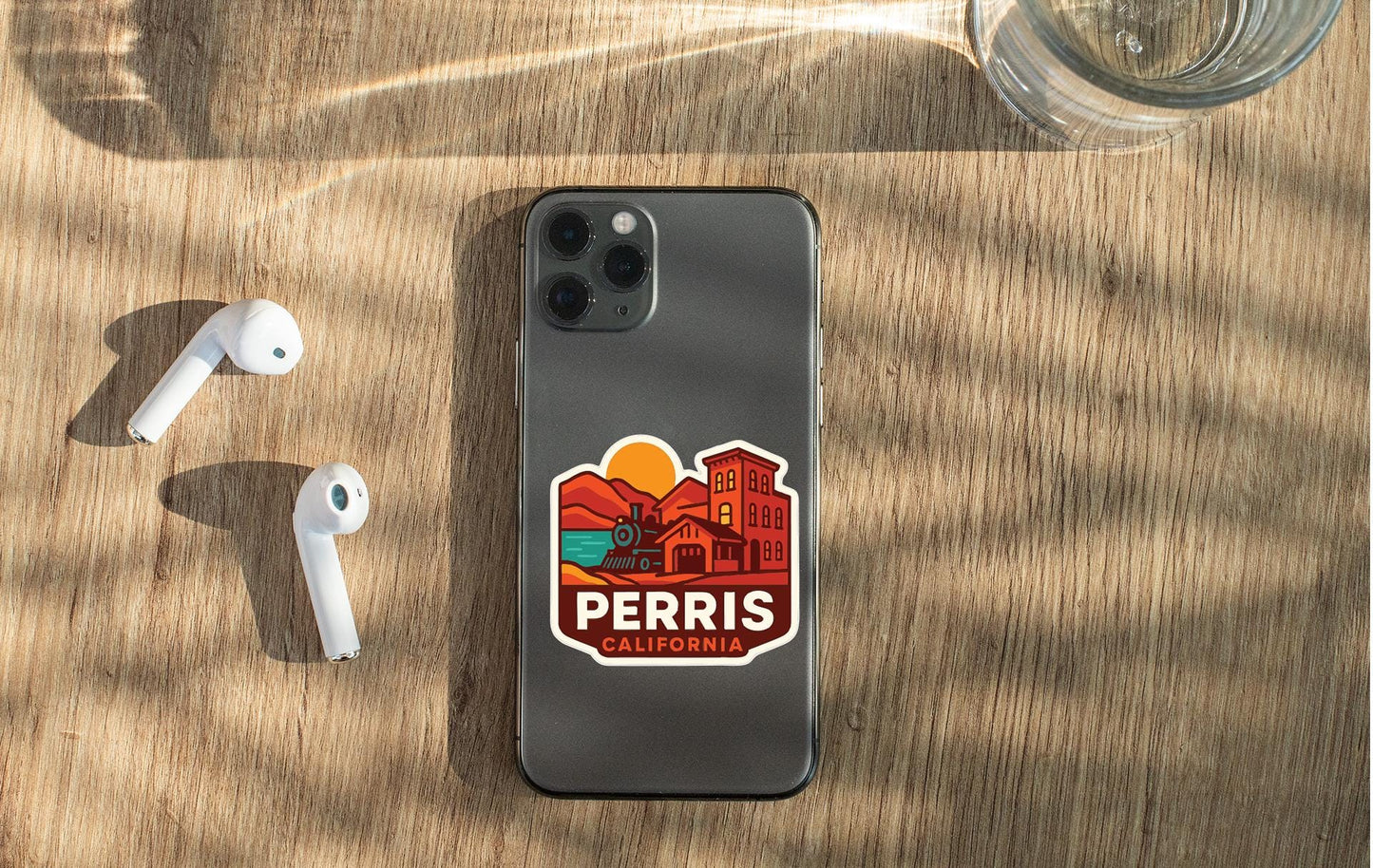 a smartphone with a sticker on the back, placed on a wooden surface alongside a pair of white earbuds and a glass of water.