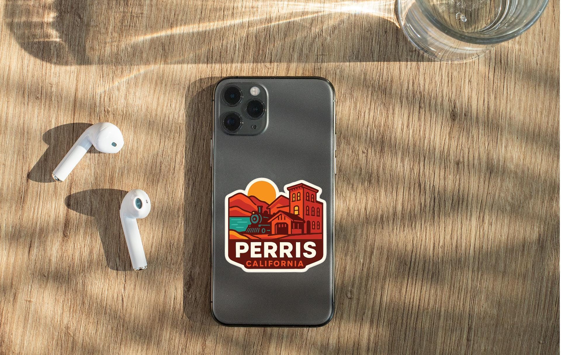 a smartphone with a sticker on the back, placed on a wooden surface alongside a pair of white earbuds and a glass of water.