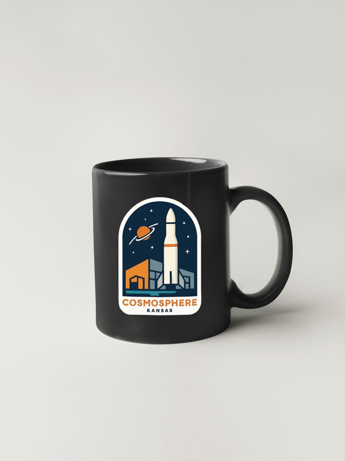 A black coffee mug with a sticker depicting a rocket ship and a city skyline, along with the text "COSMOSPHERE KANSAS".