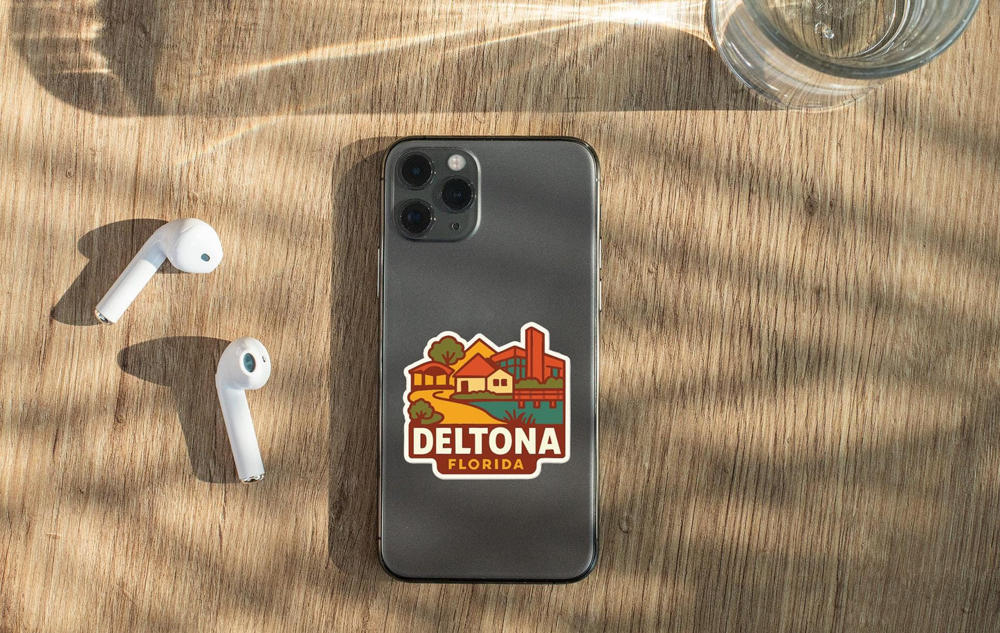 a smartphone with a sticker on the back, placed on a wooden surface alongside a pair of white earbuds and a glass of water.