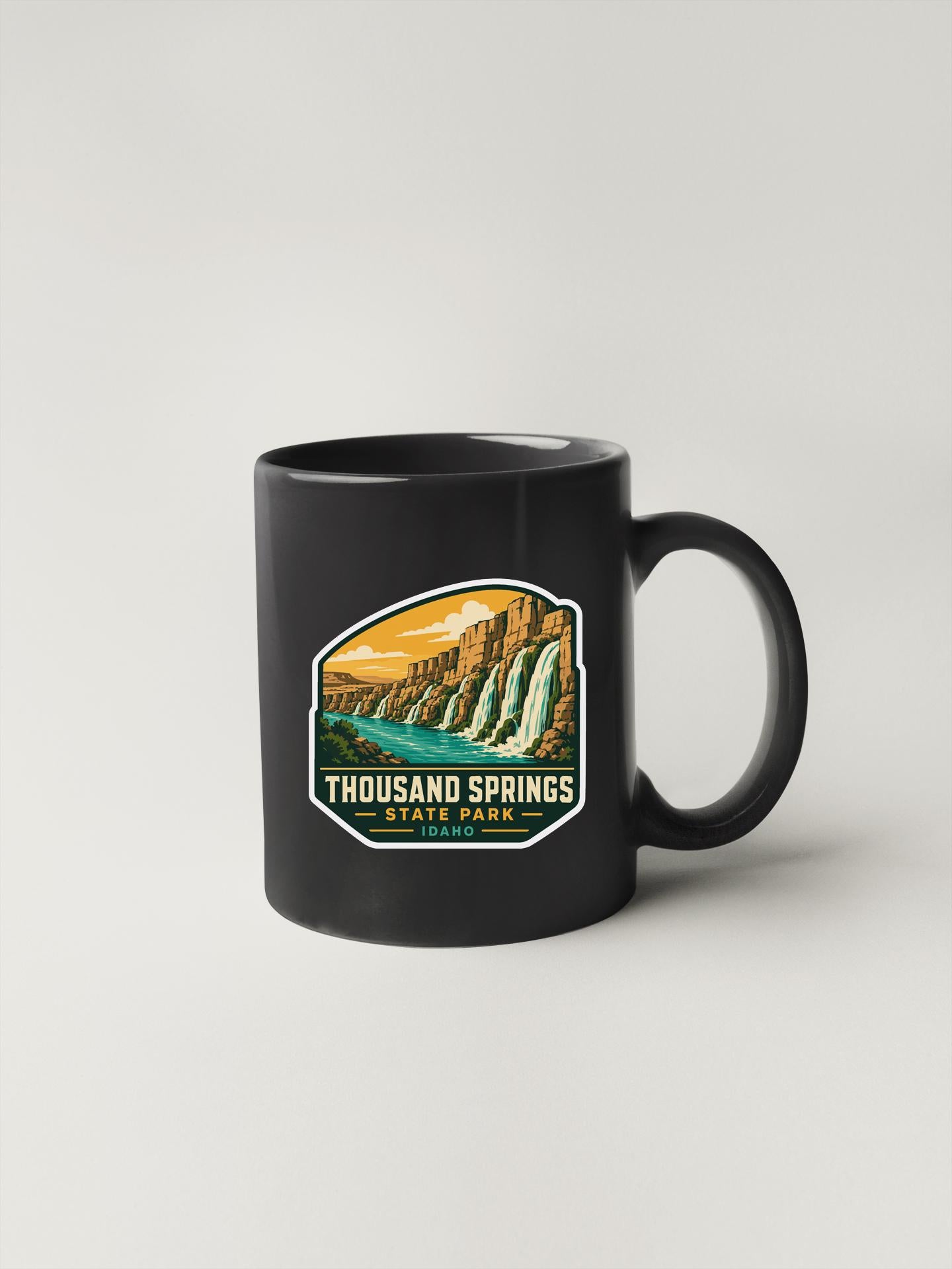 A black coffee mug with a graphic design depicting a scenic landscape featuring a waterfall and mountains.