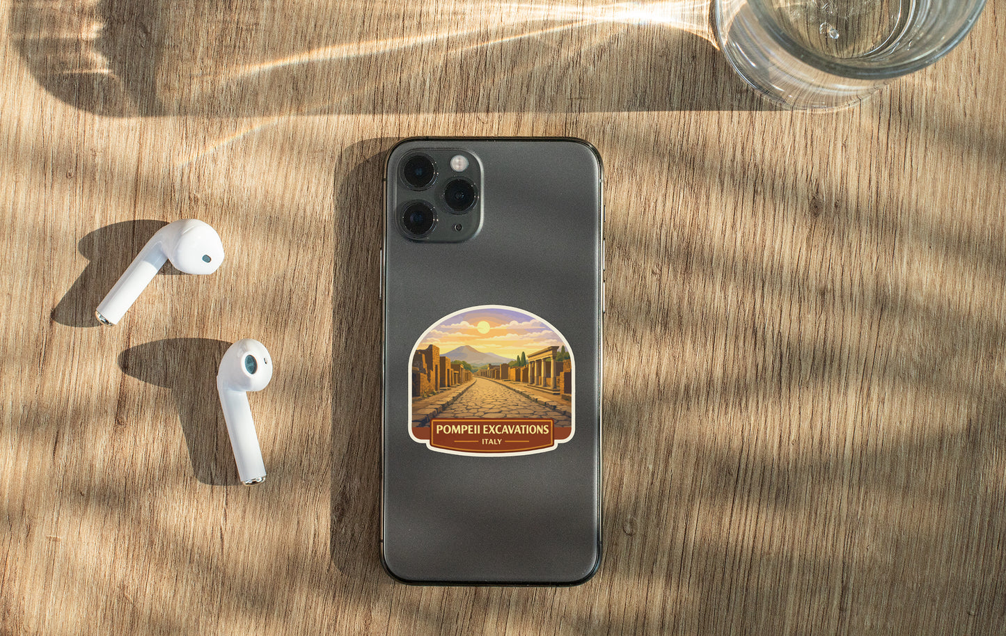 a smartphone with a sticker on the back, placed on a wooden surface alongside a pair of white earbuds and a glass of water.