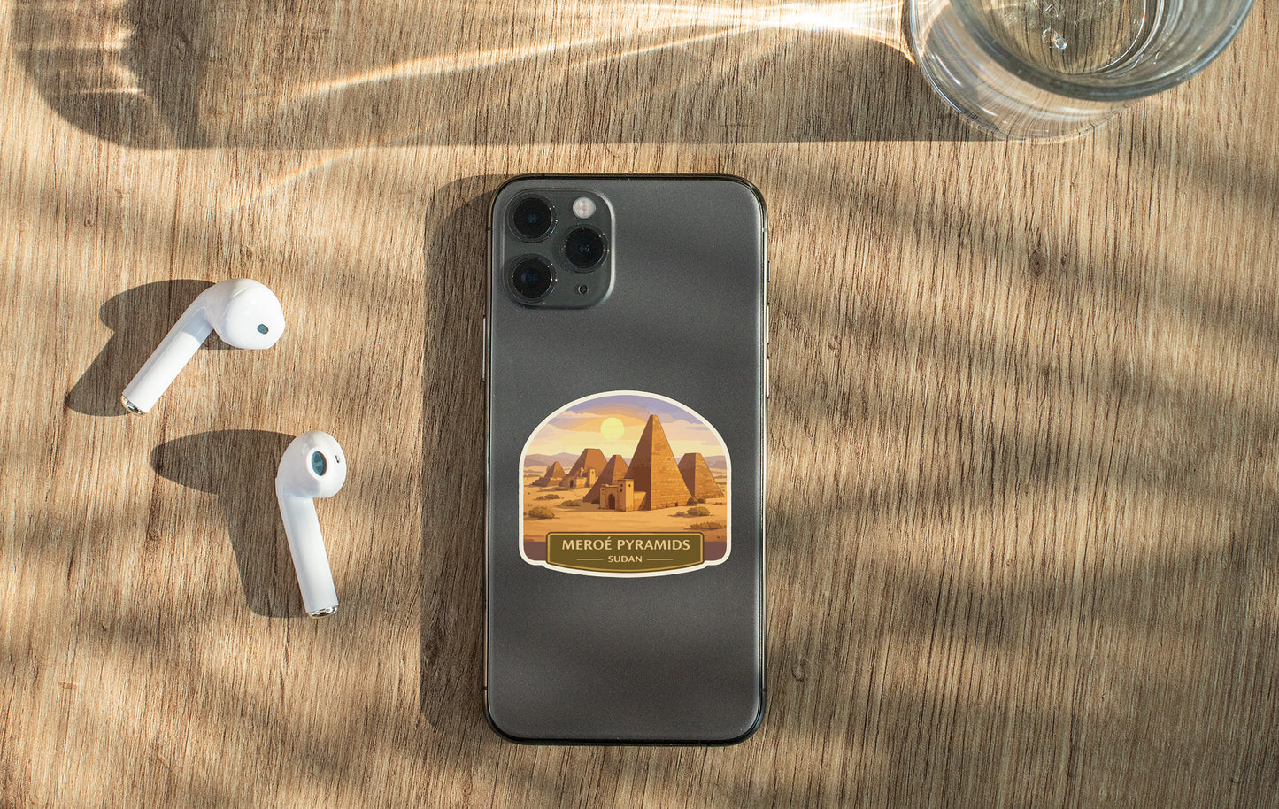 a smartphone with a sticker of the Meroe Pyramid on it, along with a pair of white earbuds and a glass of water on a wooden surface.