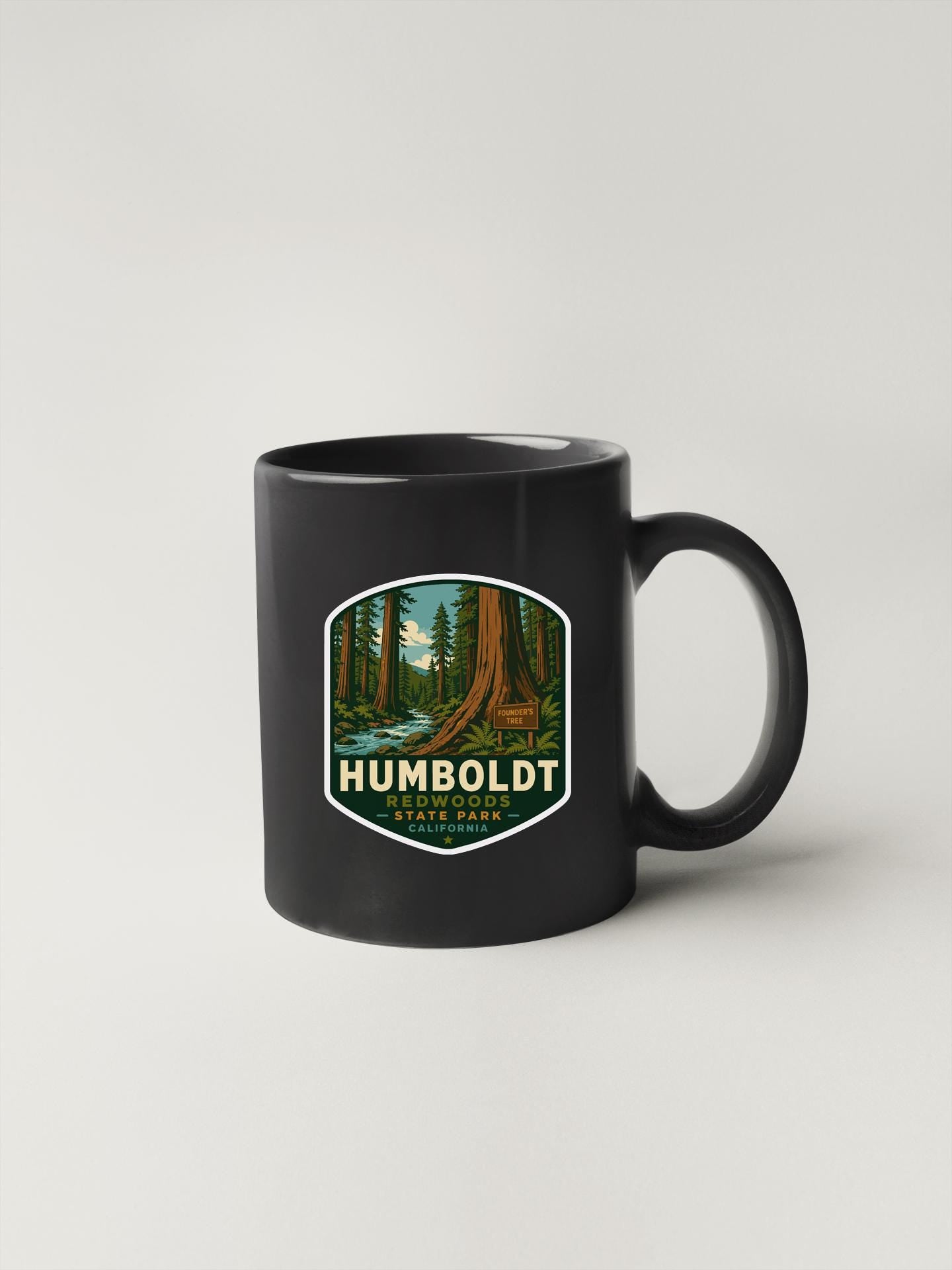 A black coffee mug with a graphic design depicting a forest scene, featuring a large tree and a stream.