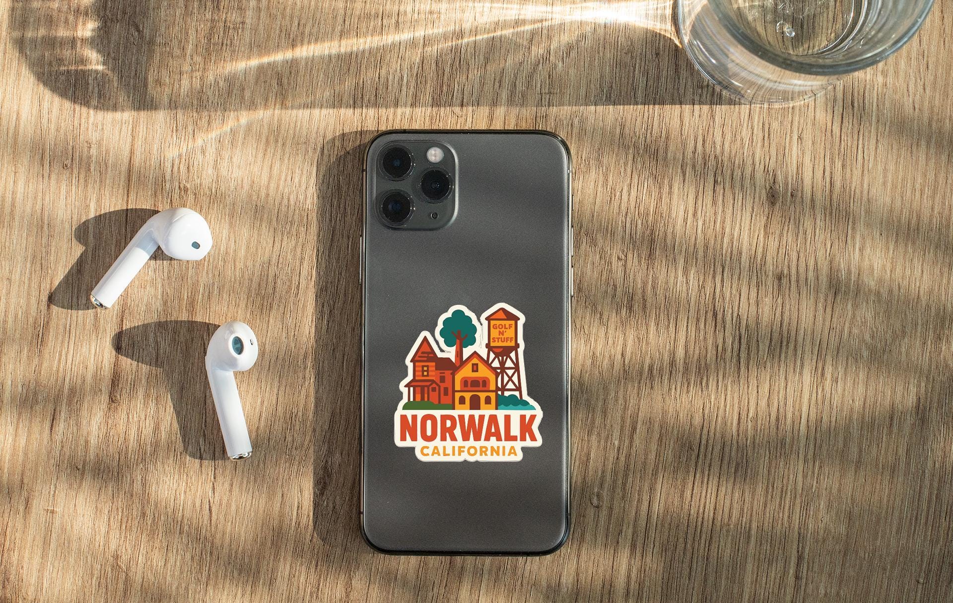 a smartphone with a sticker on the back, placed on a wooden surface alongside a pair of white earbuds and a glass of water.