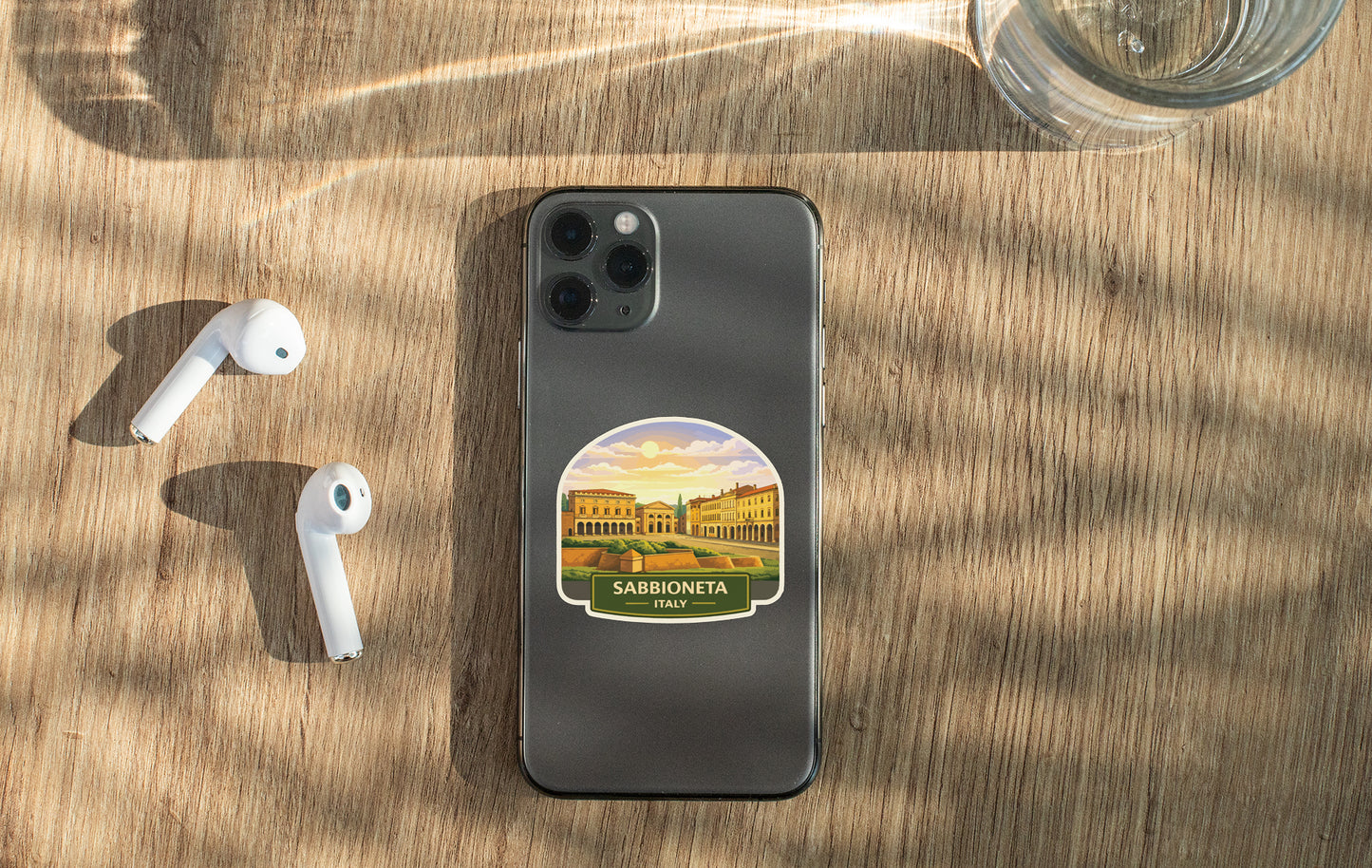 a smartphone with a sticker of a scenic view of SABIONETA, Italy, placed on a wooden surface alongside a pair of white earbuds and a glass of water.